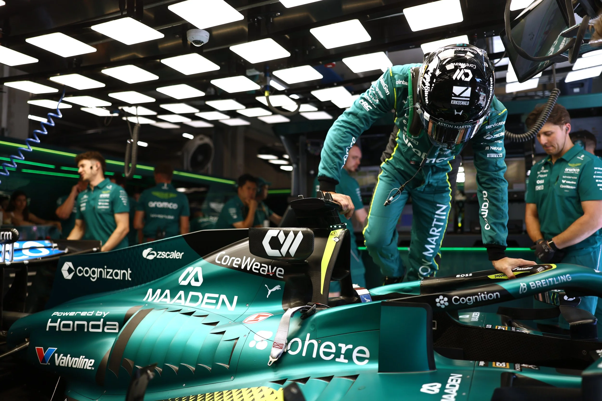 F1 team members in teal uniforms preparing a racing car in a garage, with one team member leaning over the car and wearing a helmet, while others stand nearby. The racing car displays various sponsor logos including Cognizant, Valvoline, and Honda.