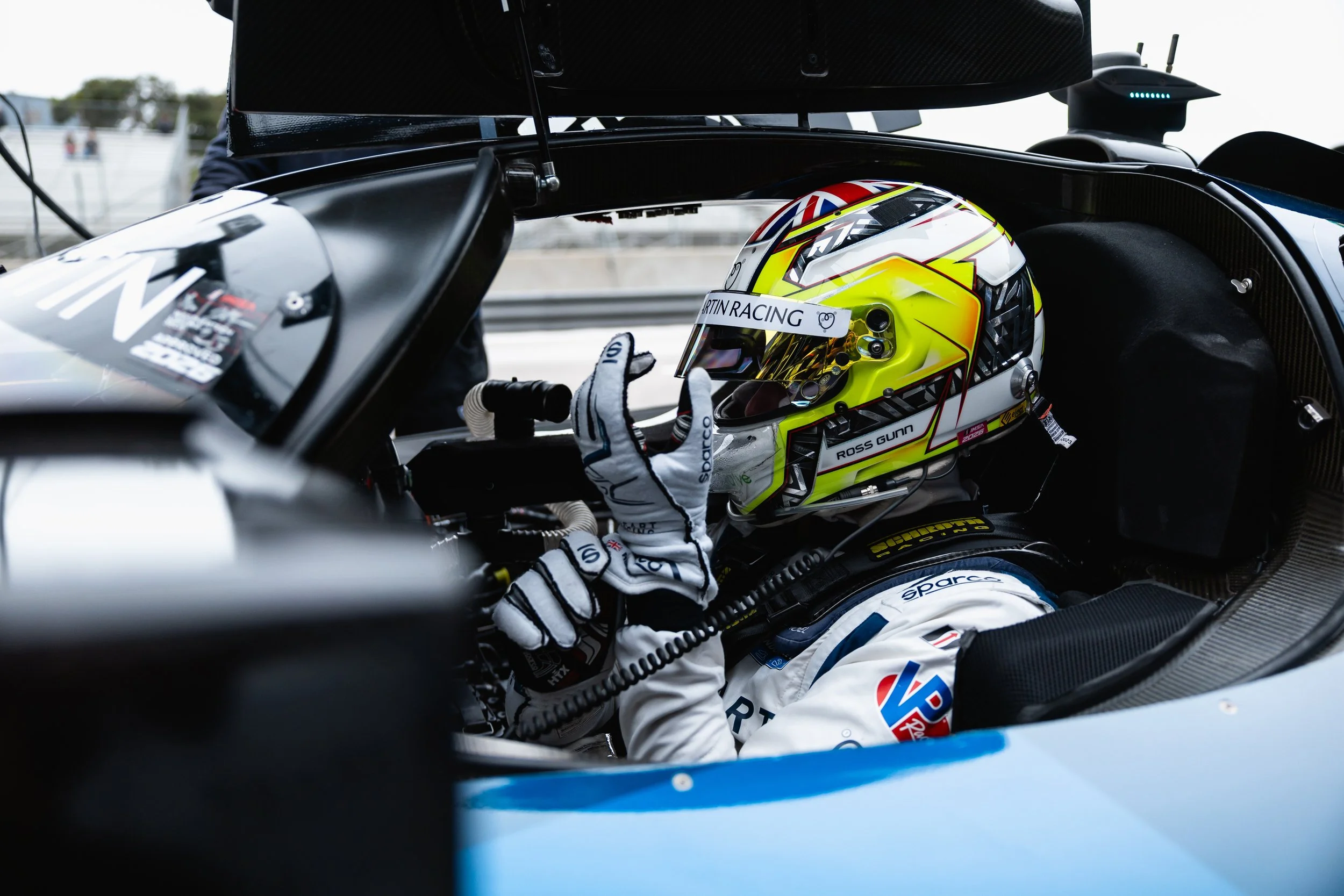 A race car driver wearing a white racing suit and gloves is seated inside a race car, adjusting their helmet, which is yellow with red and blue accents, as they prepare for a race. The car's interior is visible, with a focus on the driver's actions.