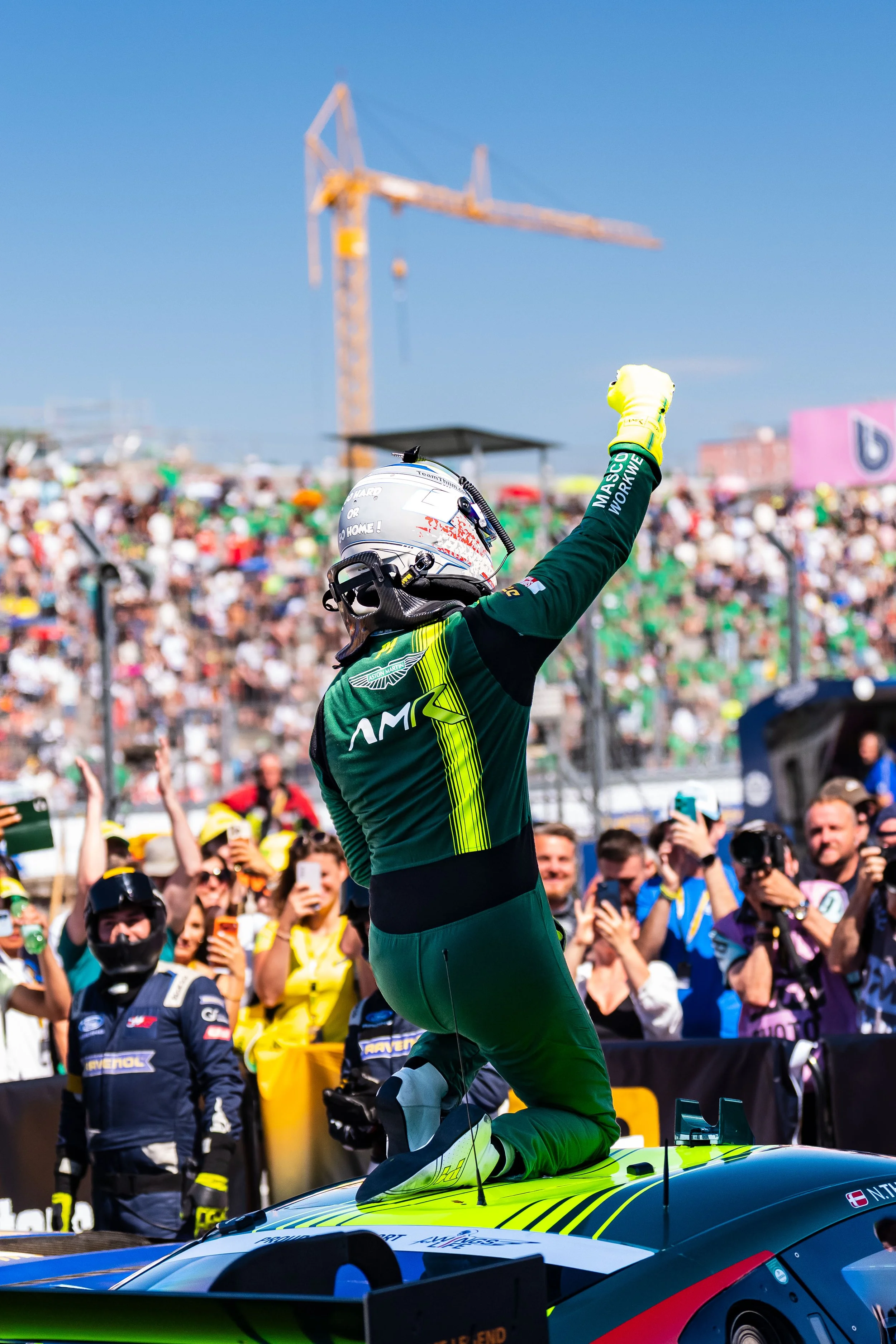 Race car driver celebrating on top of his car as crowd cheers and takes photos at racing event.