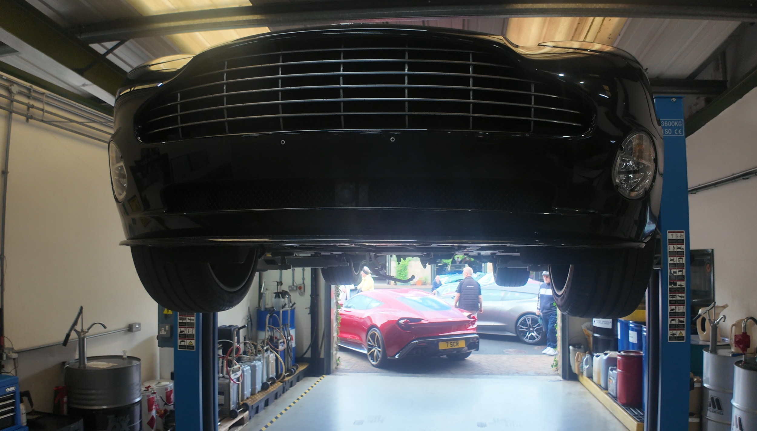 Black car lifted on a hydraulic hoist inside an automotive workshop, view from below with other cars and people outside visible through the open garage door.