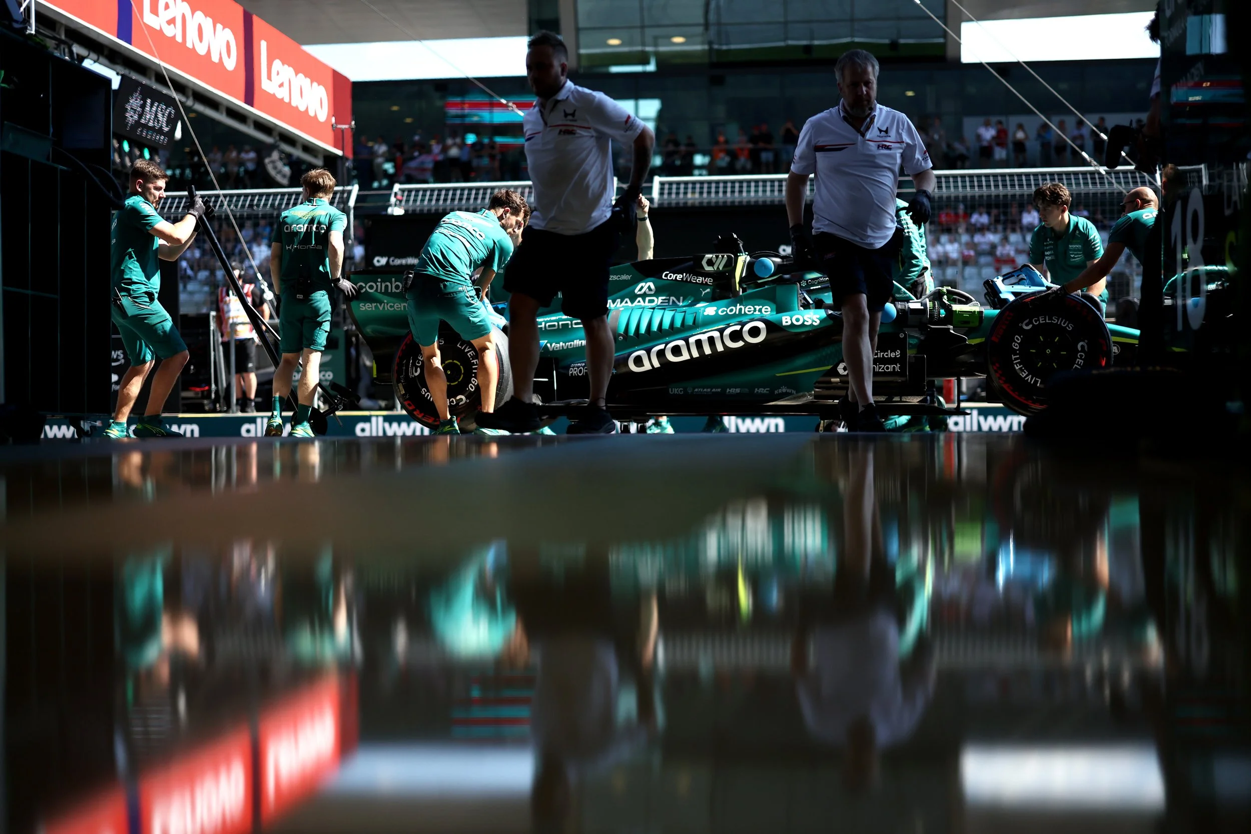Formula 1 race car being prepared in the pit lane with team members working around it, under a bridge at a racing event.