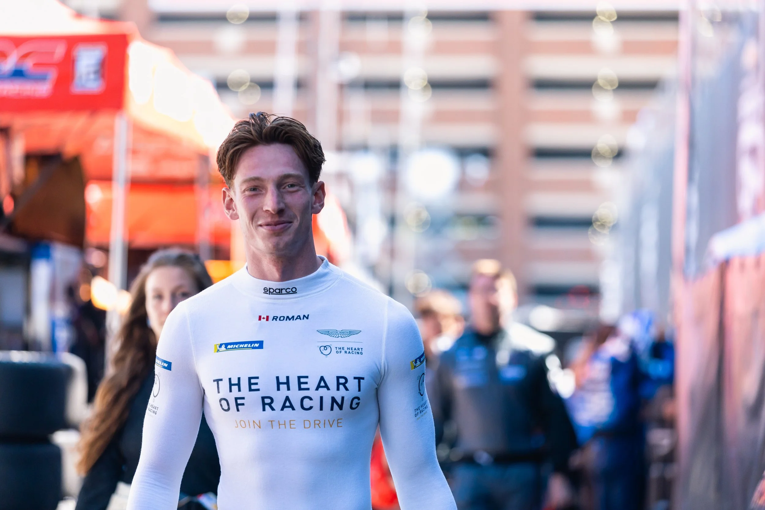 A young man in a racing suit with the name Roman, smiling, walking outdoors at a racing event with people and tents in the background.