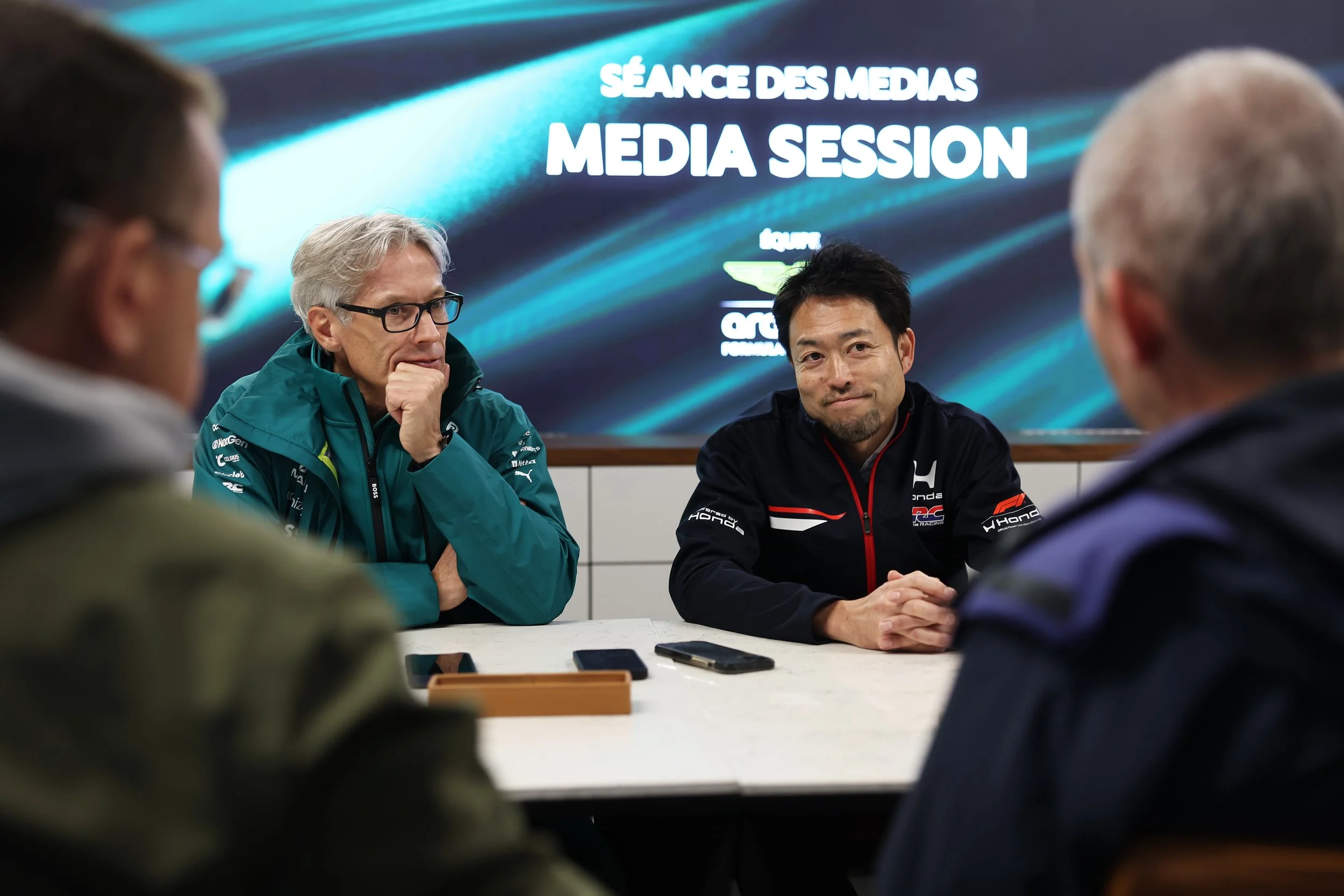 A media session with five men at a table, two in the foreground with their backs to the camera and three in the background, two of whom are facing forward. The background displays a large screen with the words 'MEDIA SESSION' and some logos or sponsors.