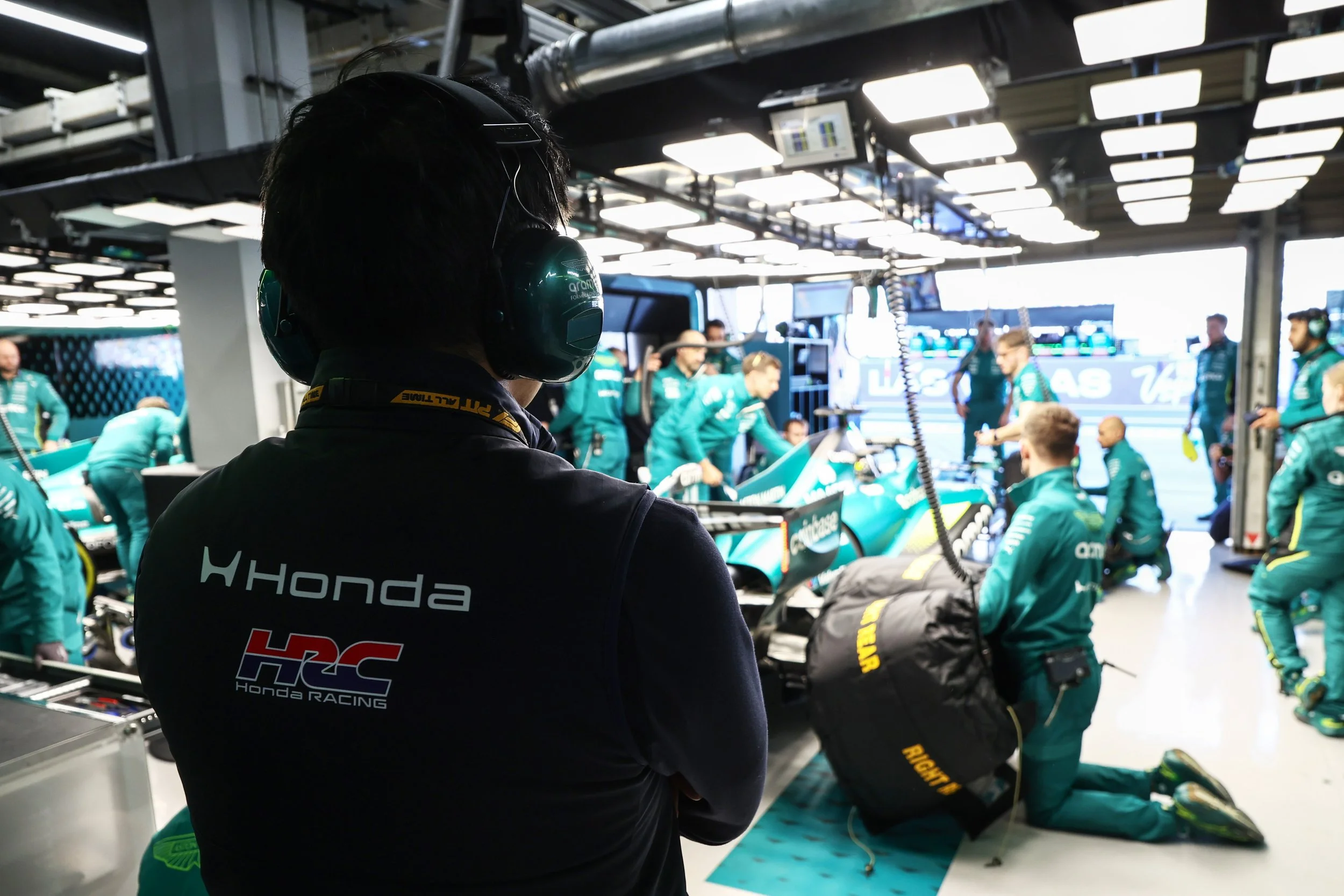 Race car team working in the garage with a team member wearing a Honda Racing shirt and headphones, team members preparing race car, racing equipment, garage lights, and monitors in the background.