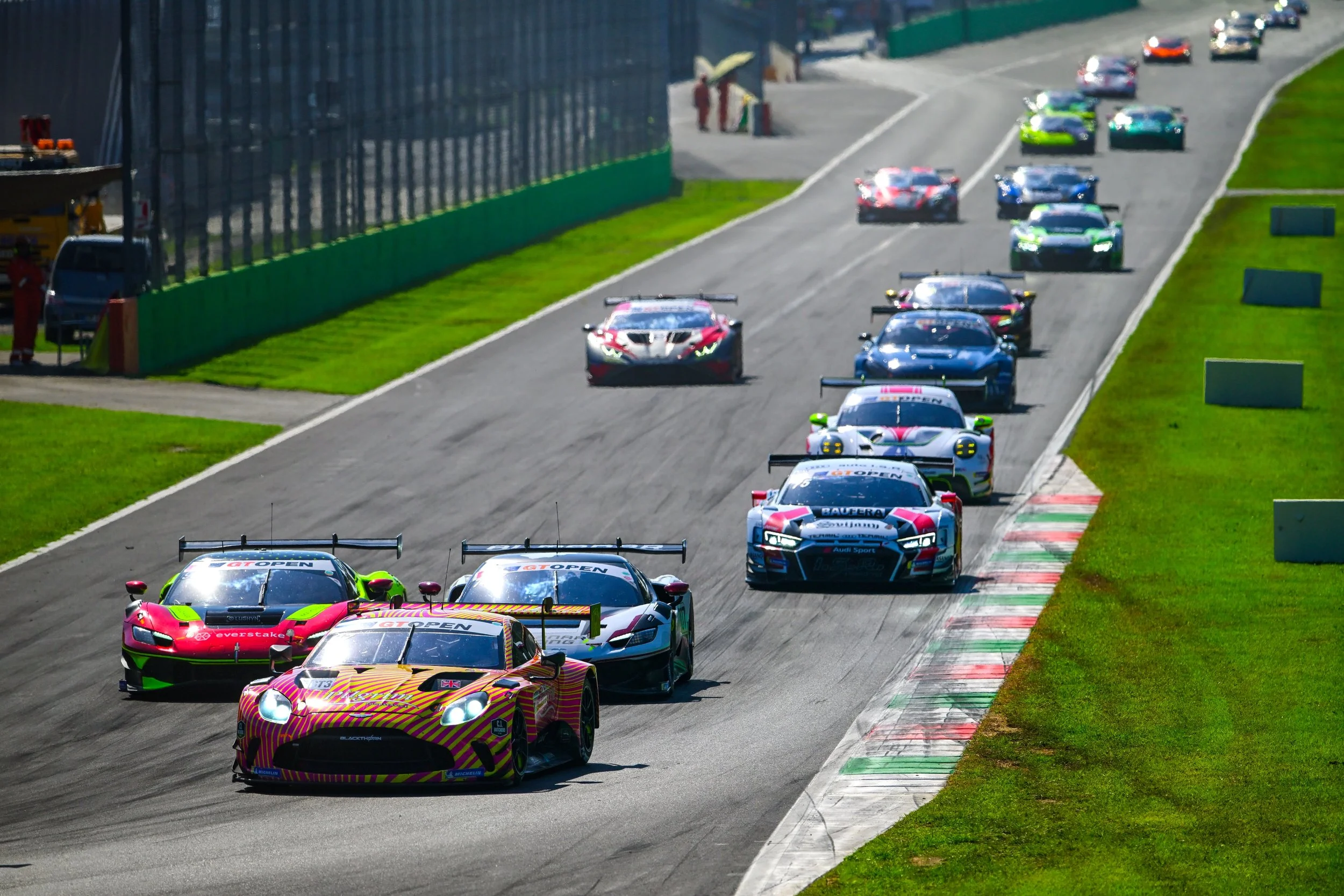A lineup of race cars on a race track ready to start a race, with some crew members and officials nearby.