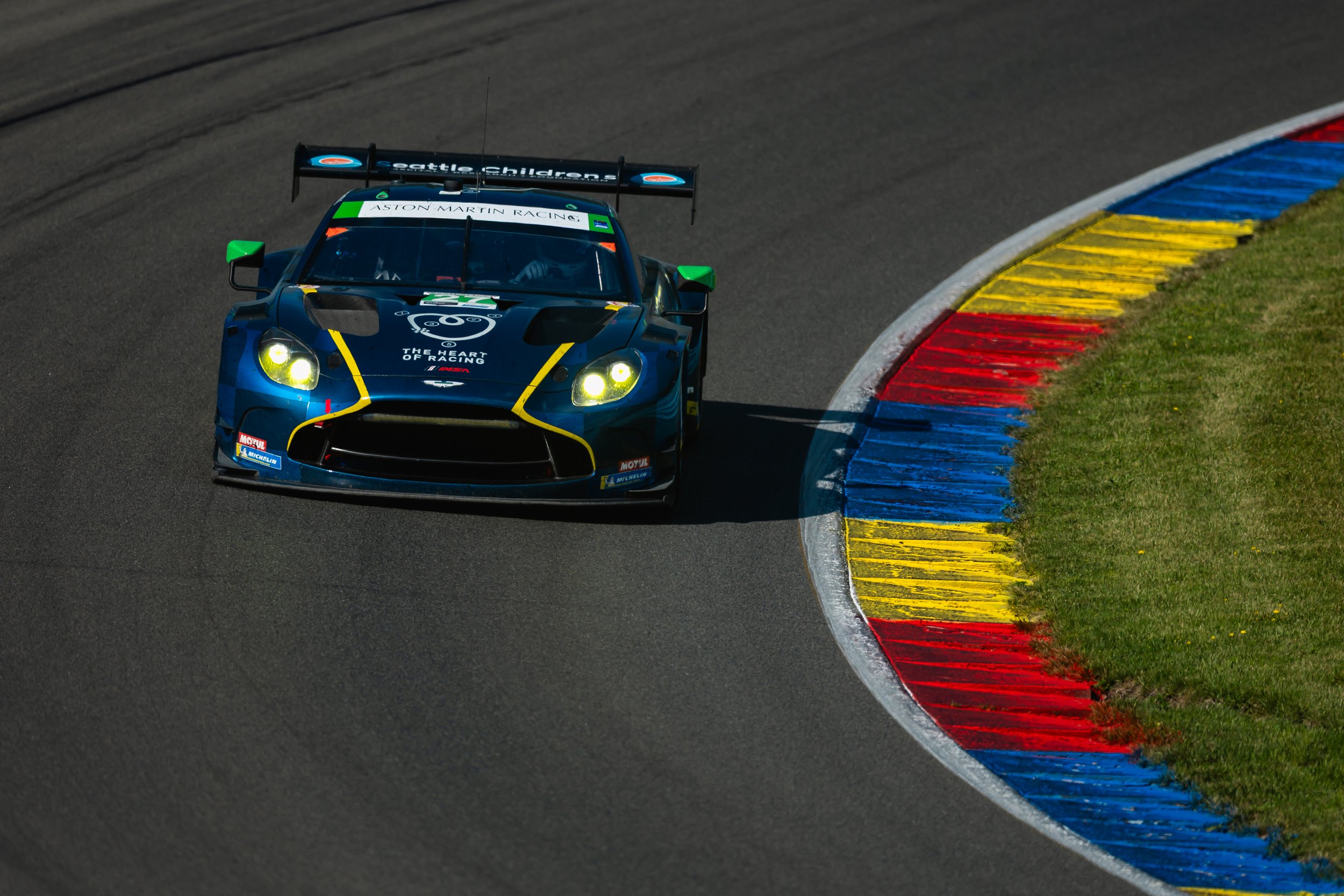 A race car on a track, navigating a sharp turn with blue, yellow, and red curbing, surrounded by a grassy area.