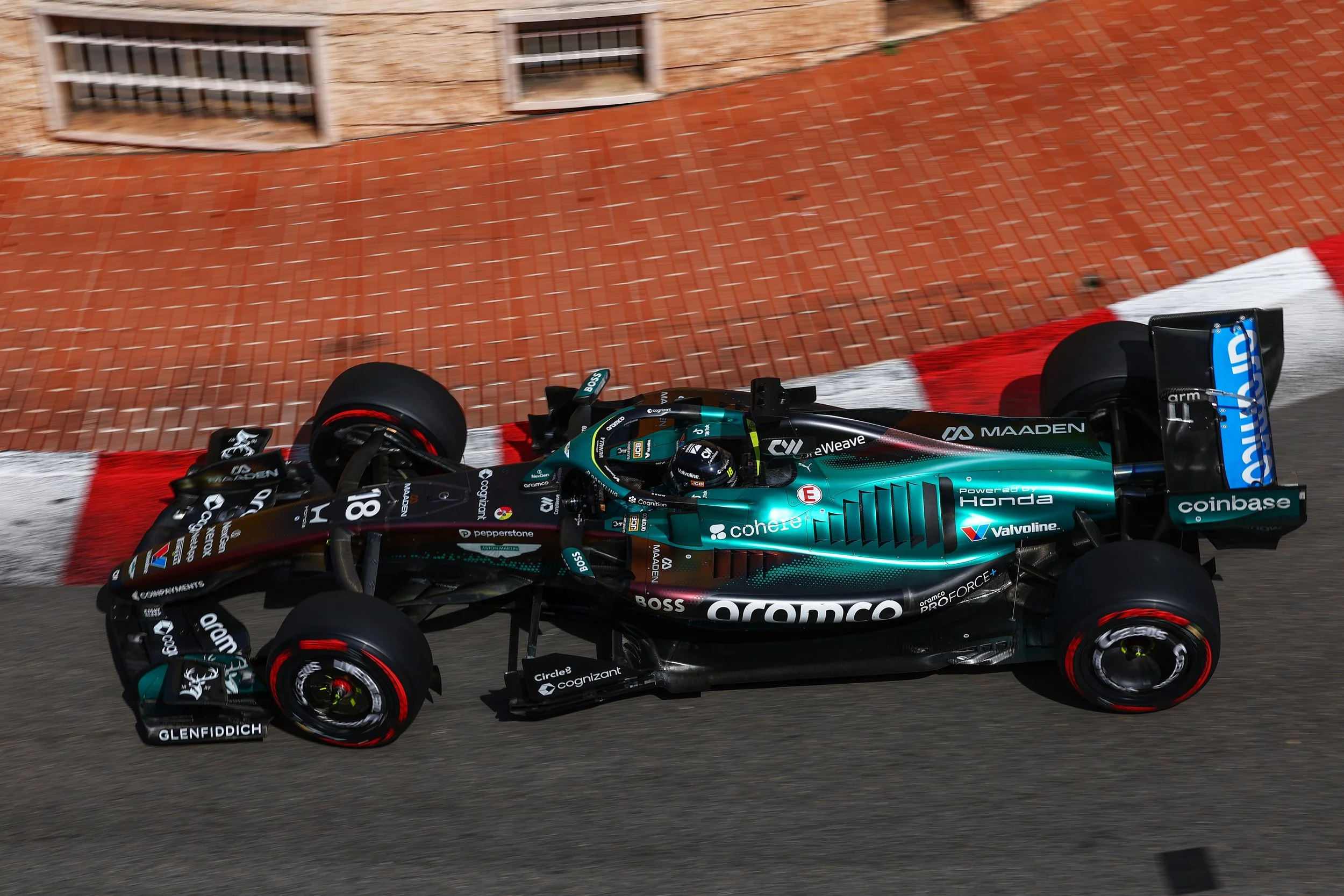 Close-up of the special livery AMR26 Aston Martin Racing Formula 1 race car at Monaco with brick wall and window in the background, featuring various sponsor logos.