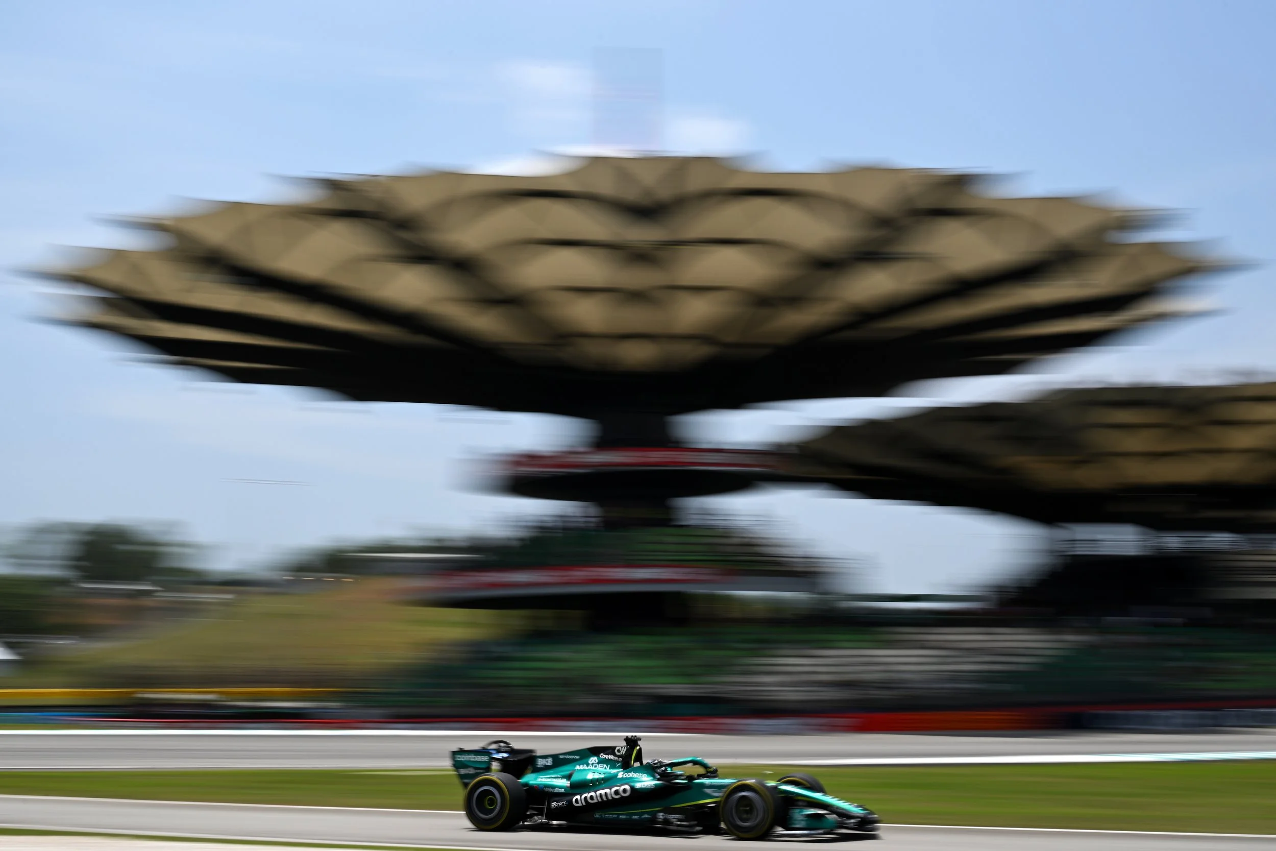 A Formula 1 race car speeding on a race track with the blurred image of a large structure or sculpture in the background and a blue sky overhead.