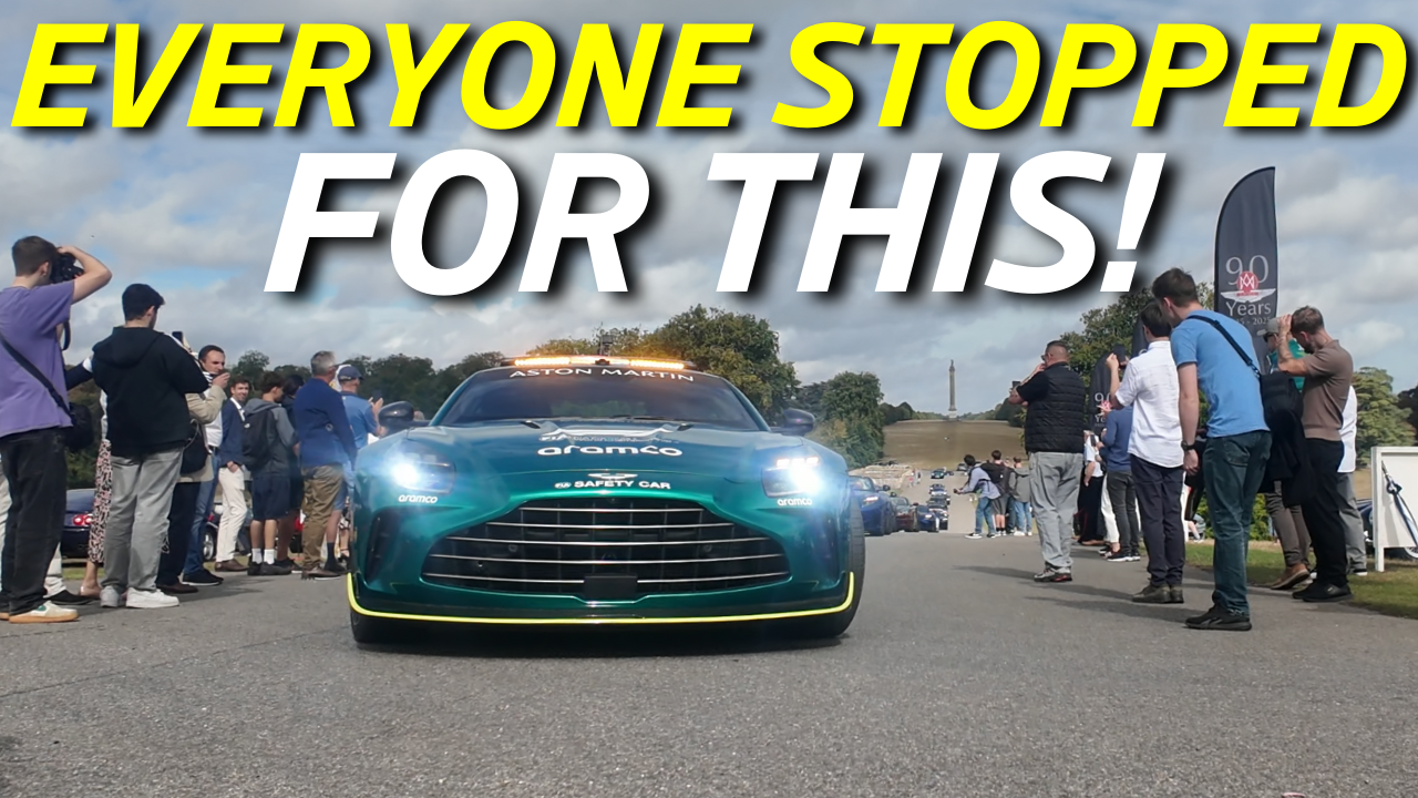An FIA Aston Martin Safety Car surrounded by a crowd of people at an outdoor event, with a cloudy sky and a monument in the background.