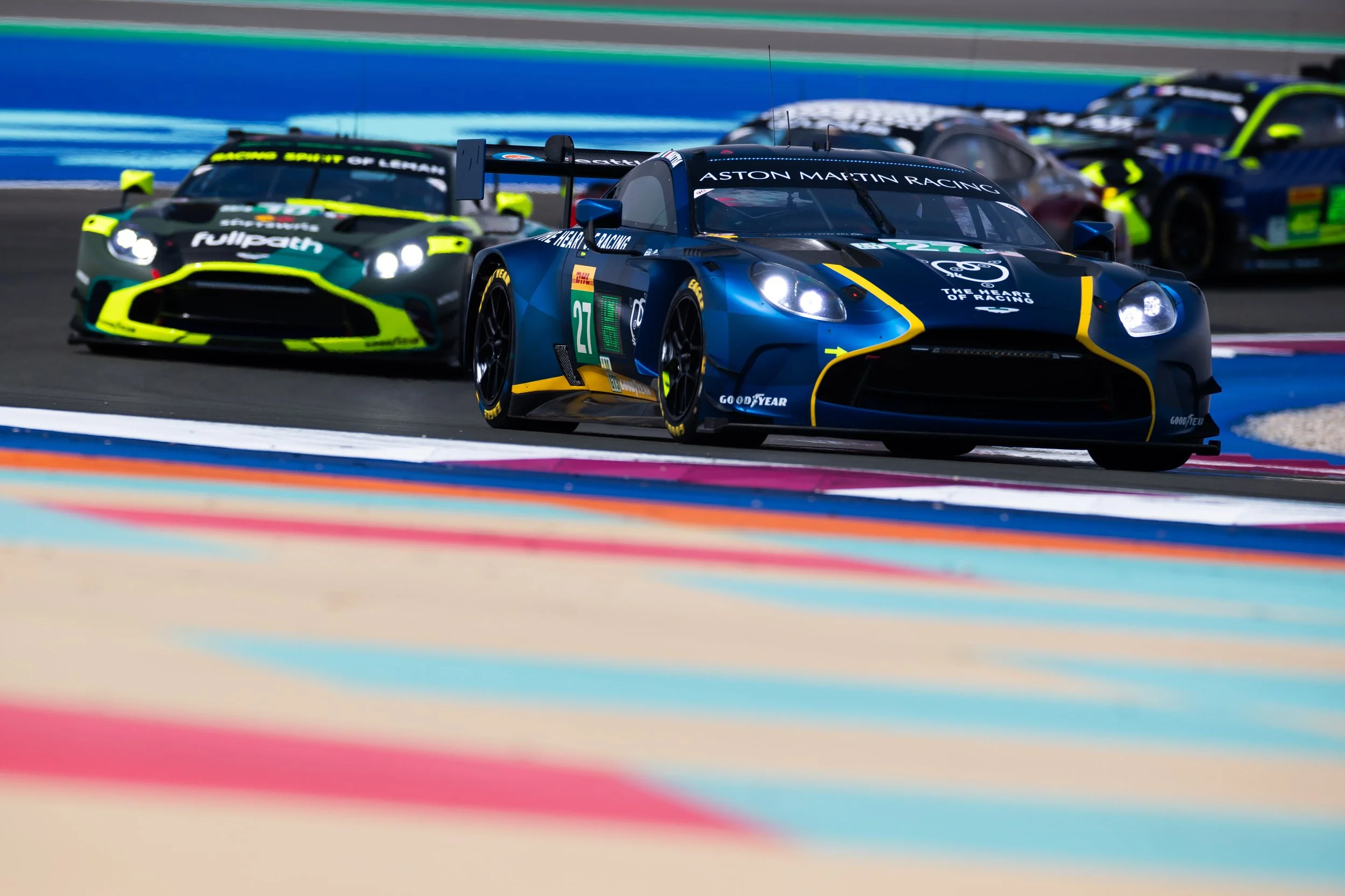 Multiple race cars on a race track, with the car in the foreground being blue with yellow accents, labeled 'Aston Martin Racing,' followed by other cars in close proximity, colorful racing curbs visible on the track.