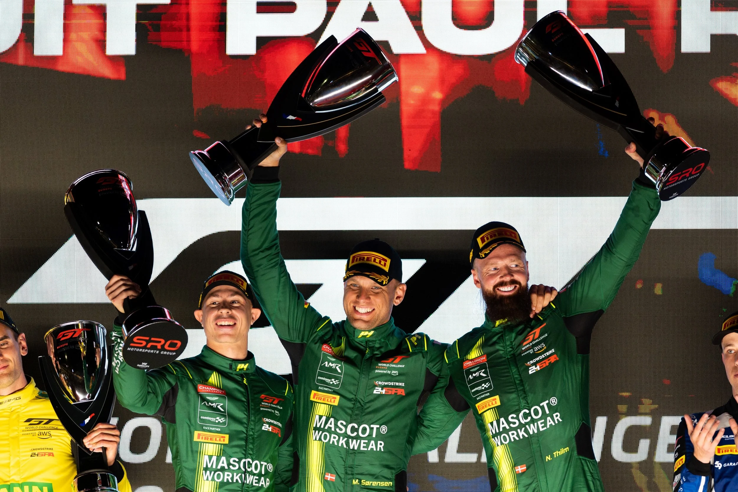Race car drivers on the podium celebrating with trophies, wearing green racing suits, holding trophies, and smiling, with a large screen and sponsor logos in the background.