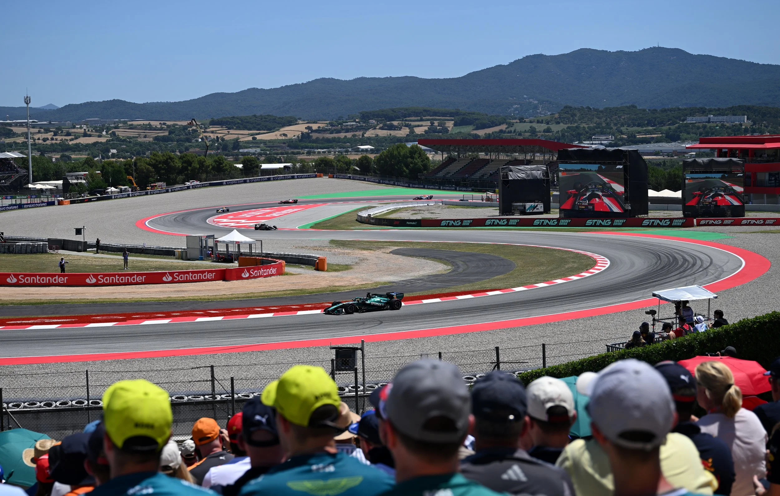 Aston Martin F1 cars, AMR26 on a track during a racing event, with spectators watching from the stands and in the foreground, mountain scenery in the background.