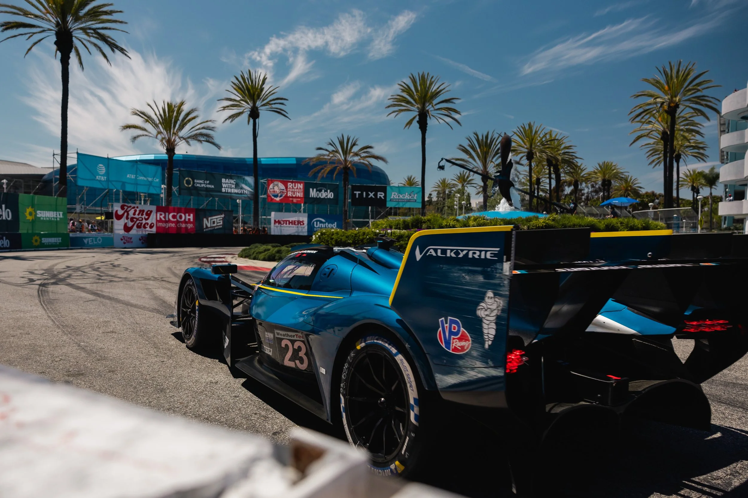 An Aston Martin Valkyrie Heart of Racing on a racing track with palm trees and a large building in the background, surrounded by advertisements and sponsor banners.