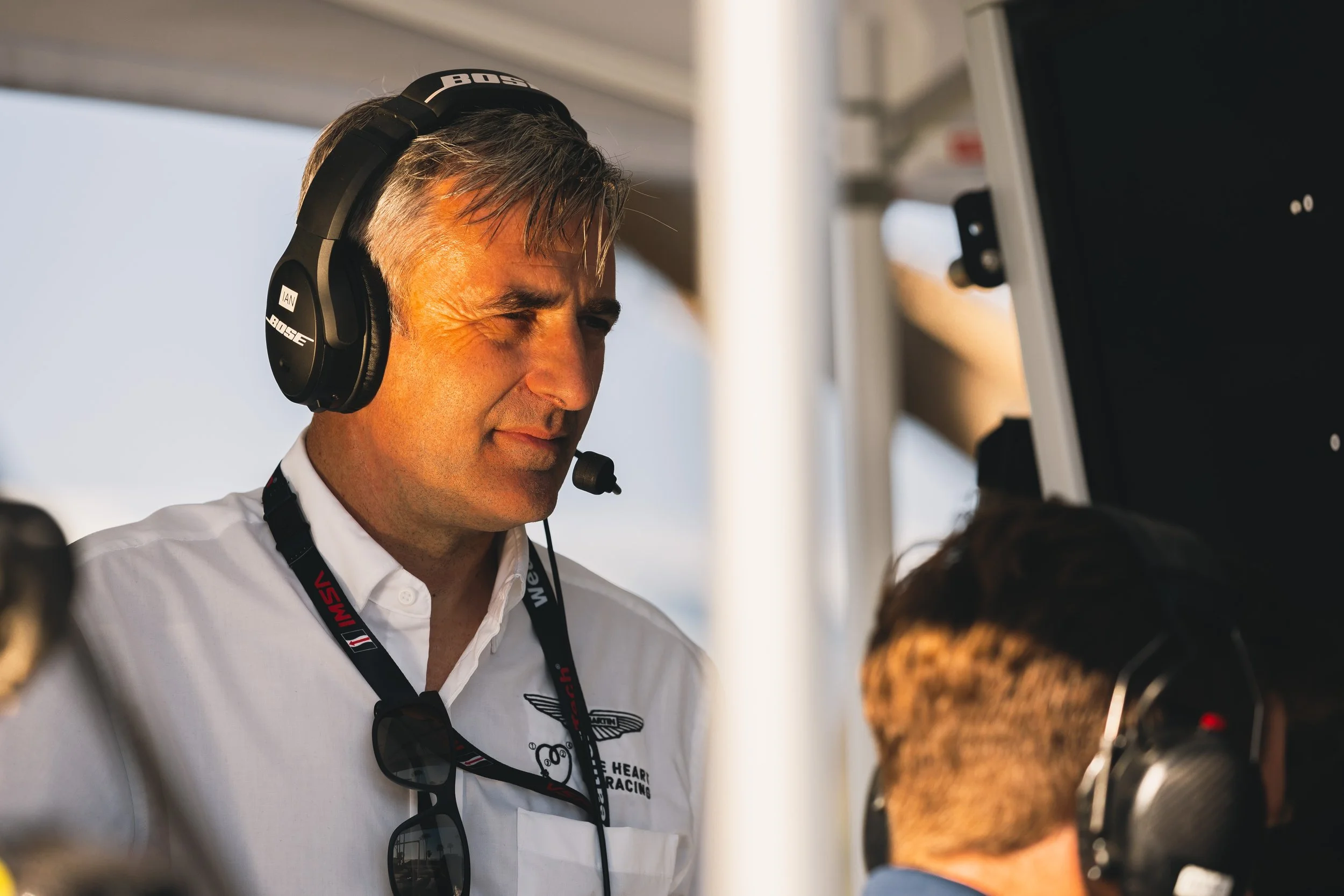 A man with gray hair wearing a white shirt, black headphones, and glasses around his neck, at a racing event, talking to a woman.