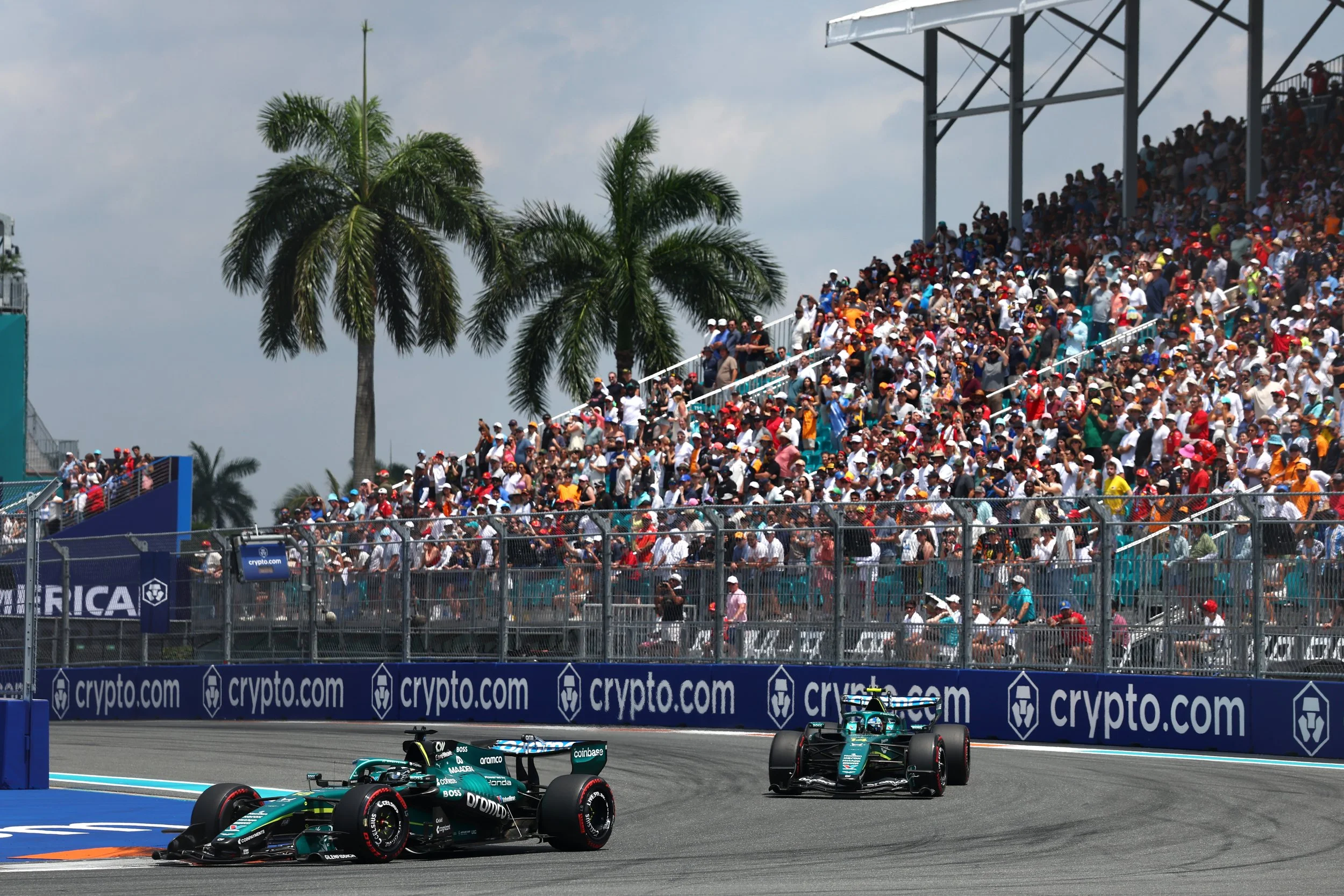 A motorsport race with Formula 1 cars on a track, held outdoors under a partly cloudy sky, with grandstands filled with spectators and palm trees in the background.