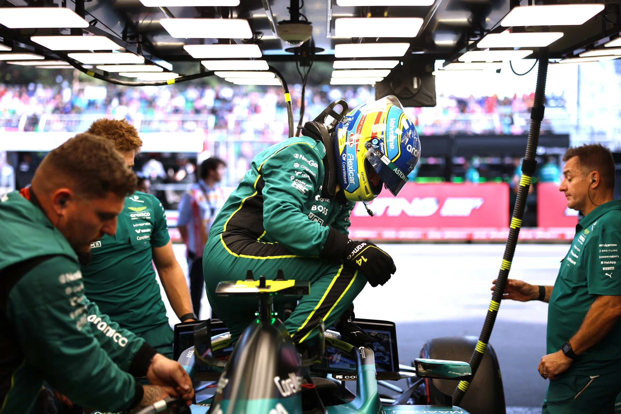 Formula 1 driver in green racing suit and helmet preparing in the pit garage, surrounded by team members working on the car.