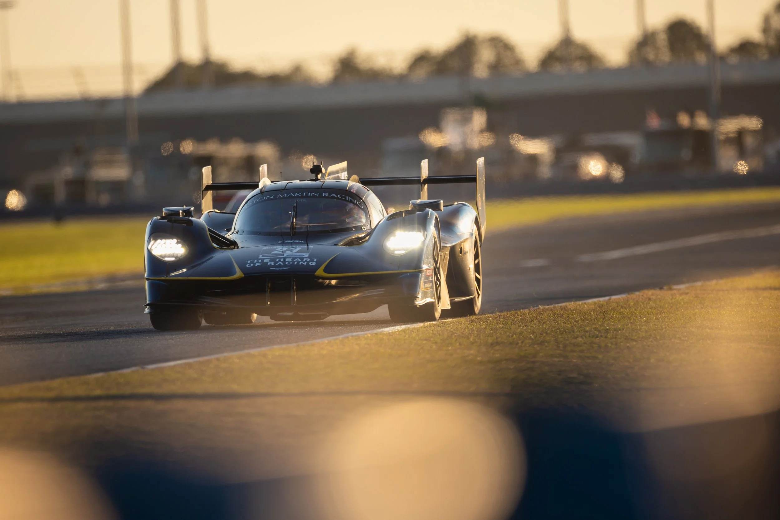 A dark blue race car with yellow accents, speeding along a racetrack during sunset, with trees and a bridge in the background.