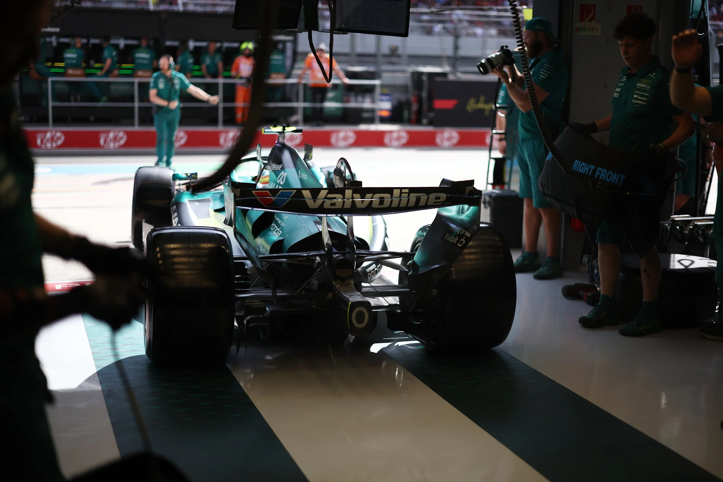 A race car inside a garage with team members working and some standing outside, with a large screen showing race-related information above.