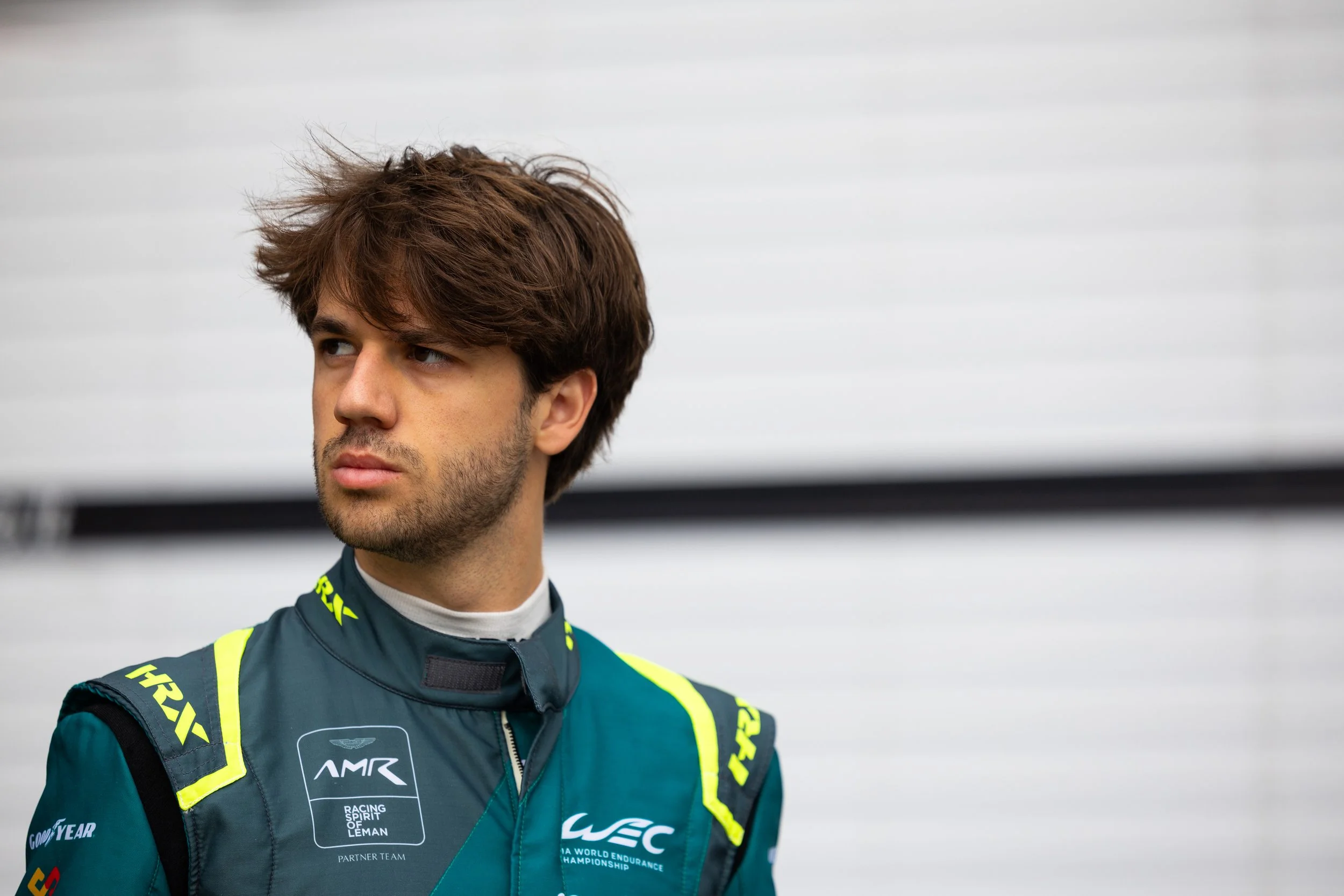 A young man with brown hair and facial hair wearing a racing suit with various sponsor logos, looking to the left with a focused expression, against a blurred neutral background.