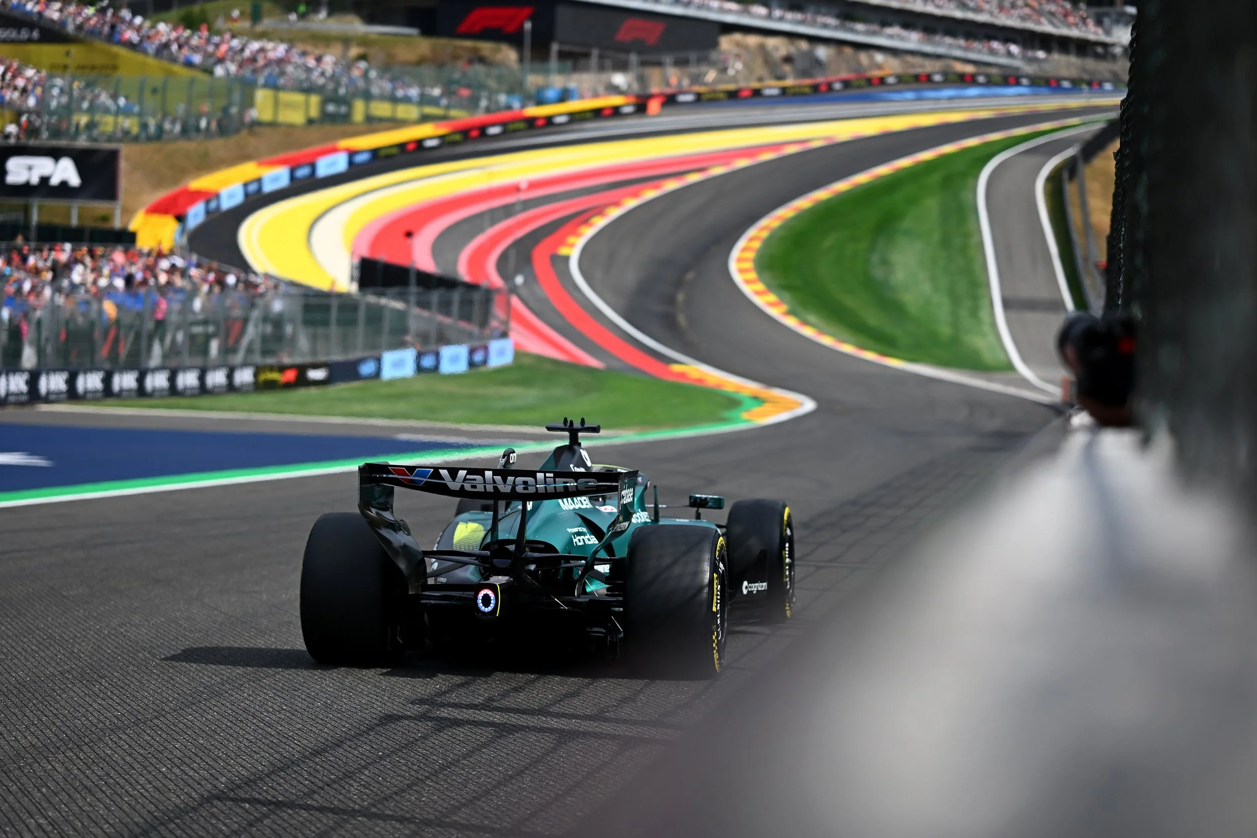 Formula 1 race car on a racing track during a race, with spectators in the stands and colorful painted curbs.