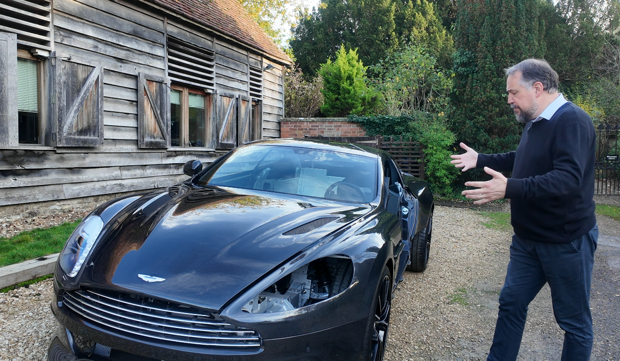A man gesturing towards a black Aston Martin sports car with some missing headlight or front panel parts, parked on a gravel driveway near a wooden house and greenery.