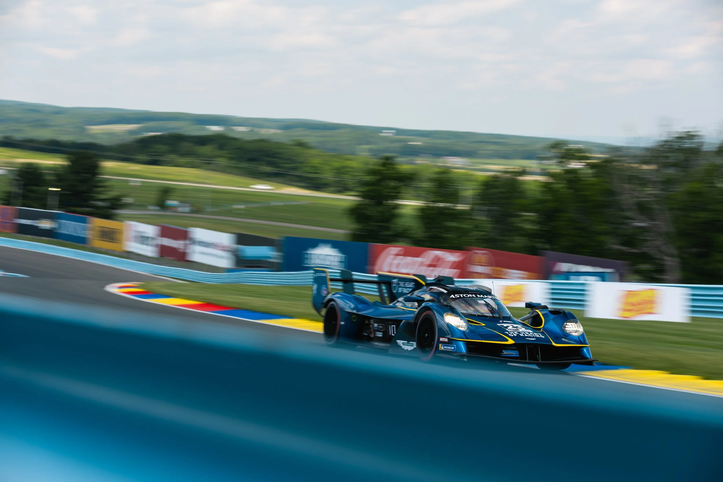 A sleek racing car on a race track, with a background of trees, hills, and a cloudy sky, blurred to emphasize speed.
