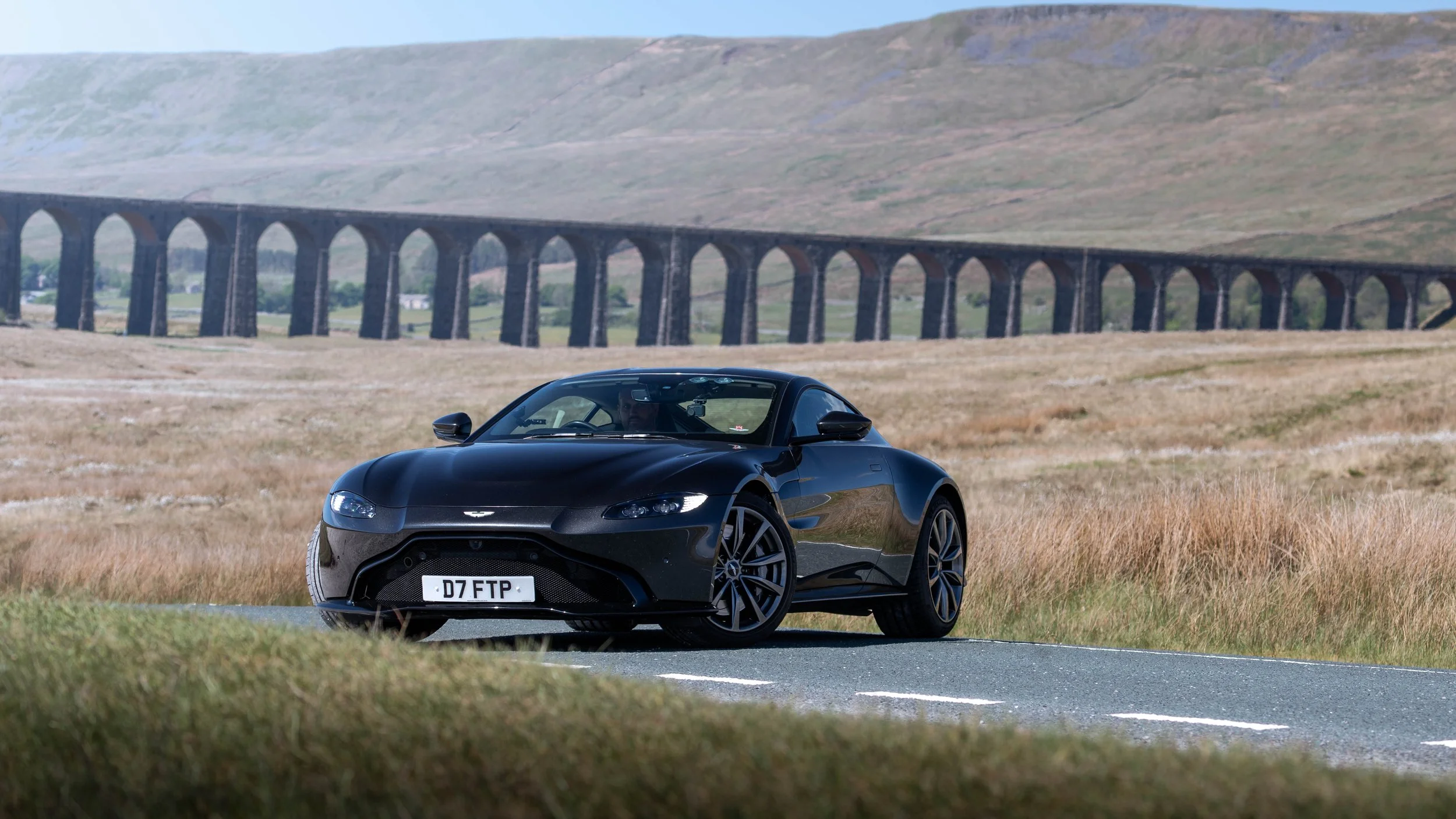 A black Aston Martin sports car parked on the side of a rural road with tall grass, with a large stone arch bridge and rolling hills in the background.