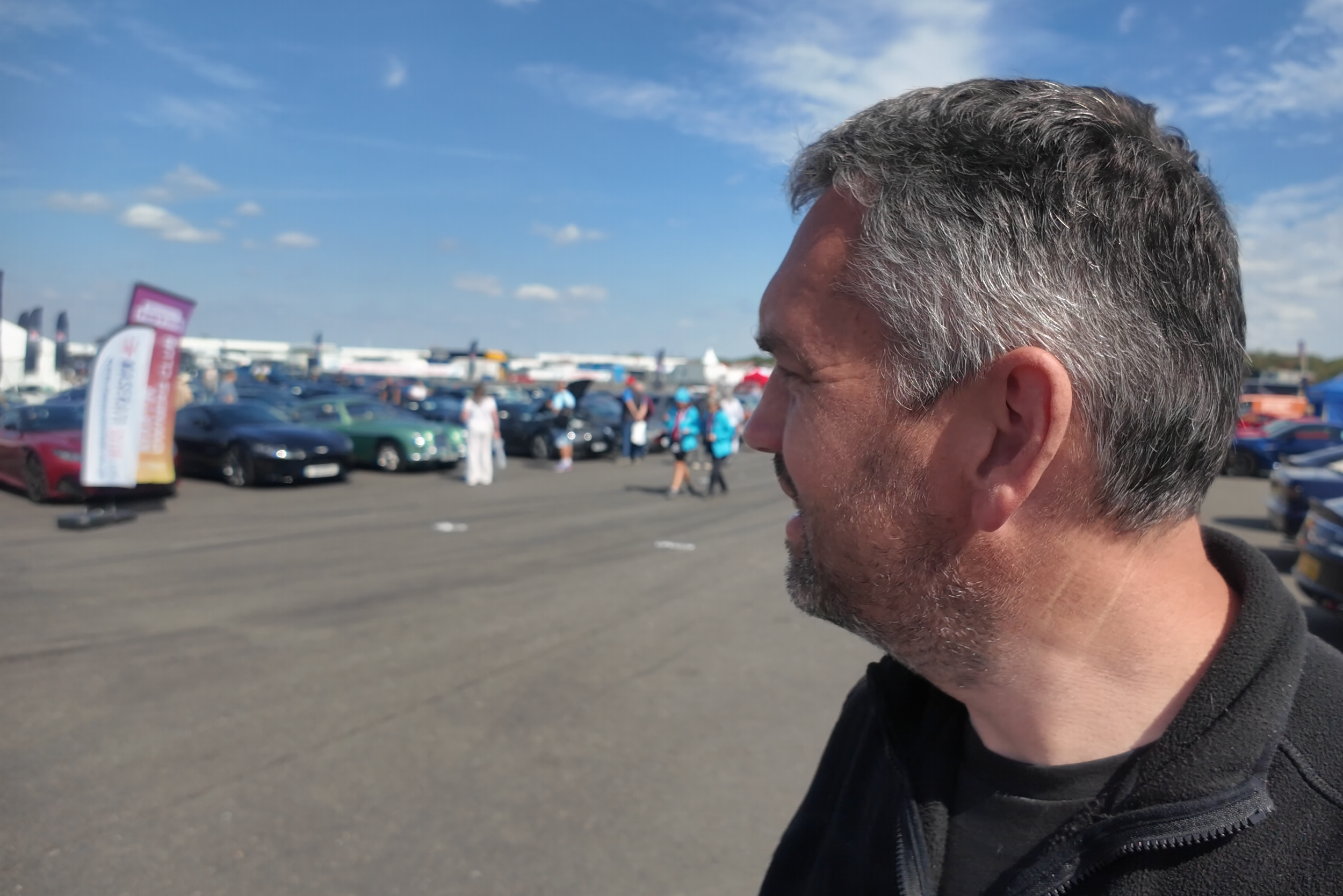 Side profile of Dan from Fuel the Passion, standing in a busy outdoor car show, with many cars and people in the background on a clear, sunny day.