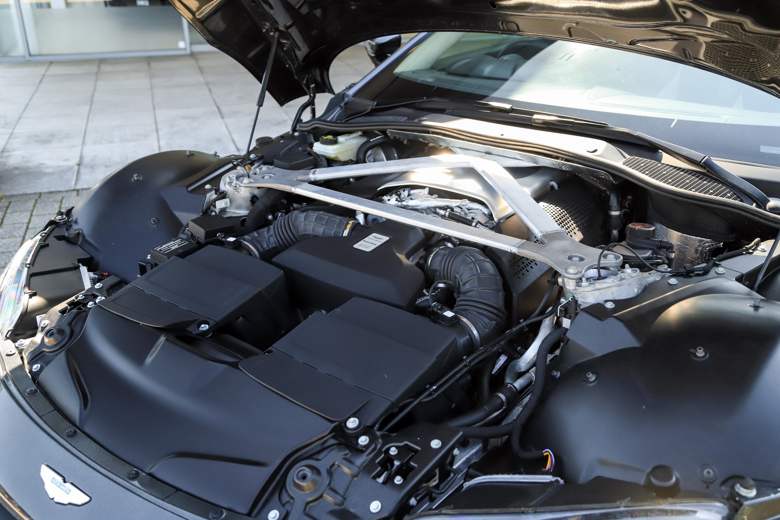 Under the hood of a black sports car, showcasing engine components, air intake, and structural supports.
