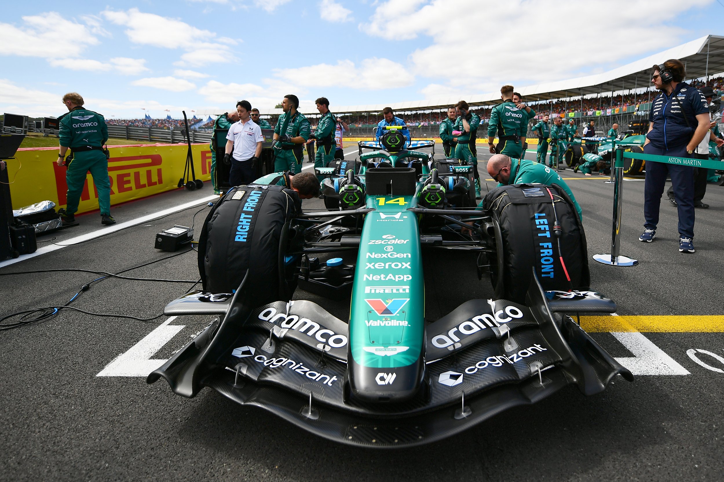 A Formula 1 race car on the grid with team members working around it during a race event on a partly cloudy day.