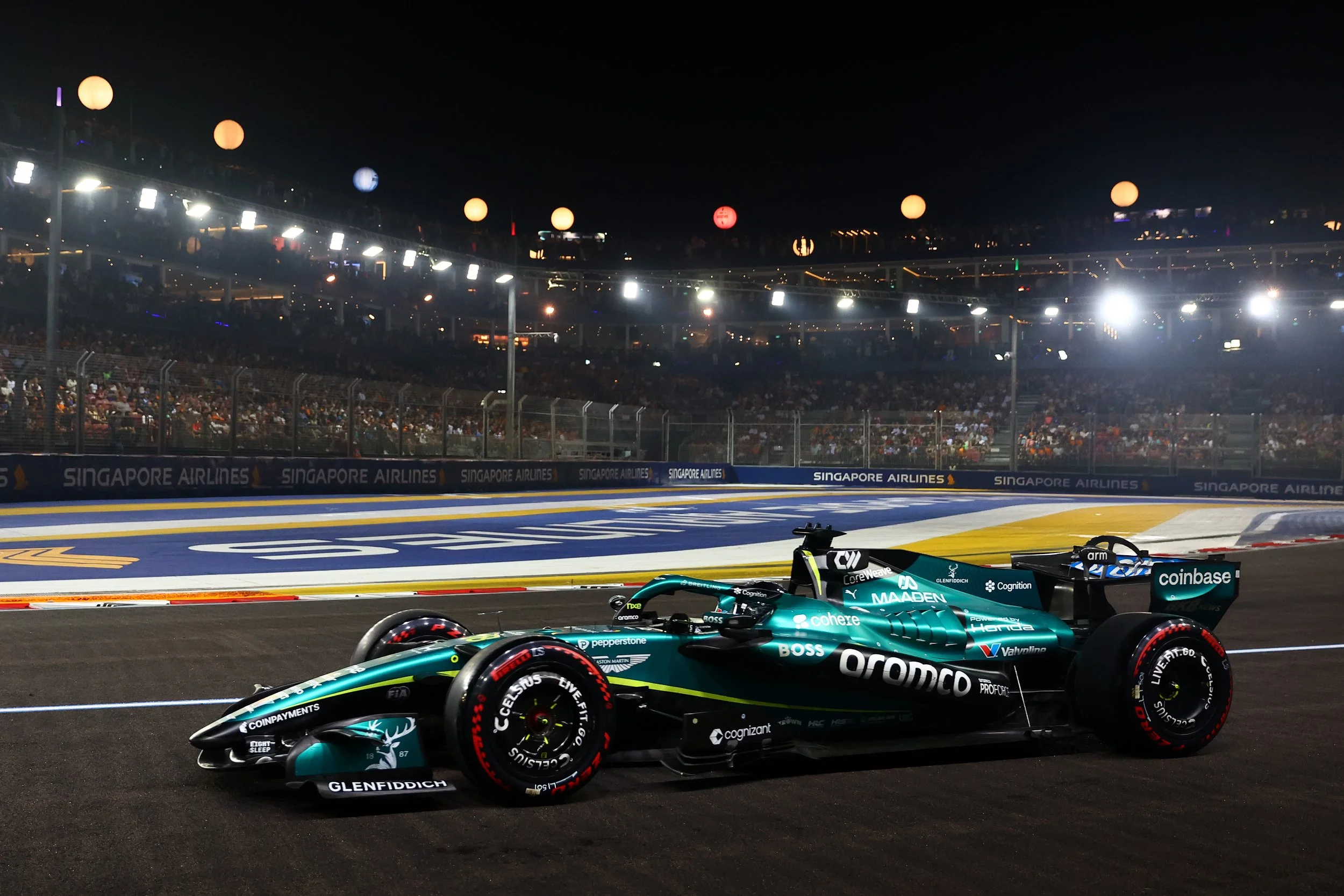 Nighttime Formula 1 race track with racing cars on the track and spectators in the grandstands, illuminated by bright lights, featuring Singapore Airlines advertising and colorful stadium lighting overhead.