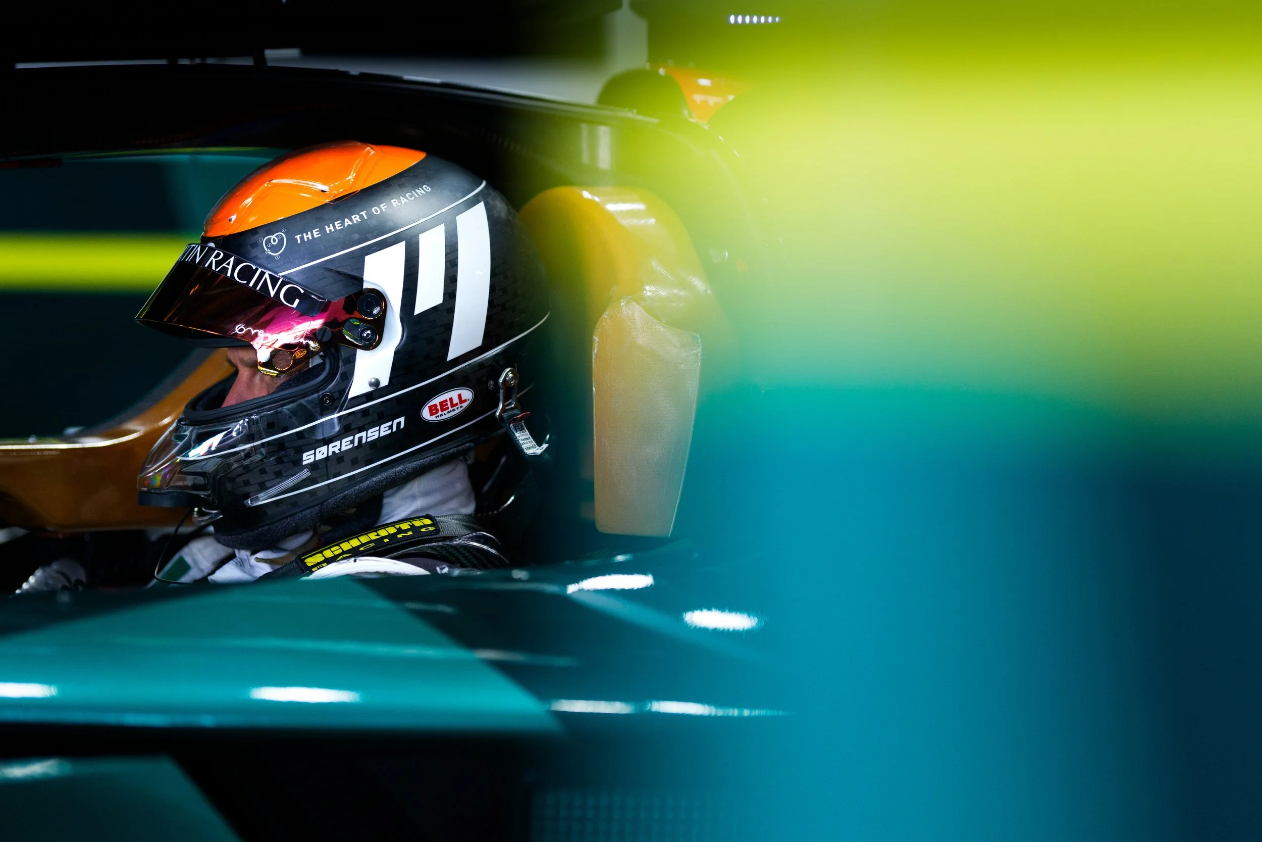 A race car driver wearing a black helmet with orange accents and sponsor logos, sitting inside a racing car. The helmet has the phrase 'THE HEART OF RACING' printed on it. The car's interior and windshield partially cover the driver.
