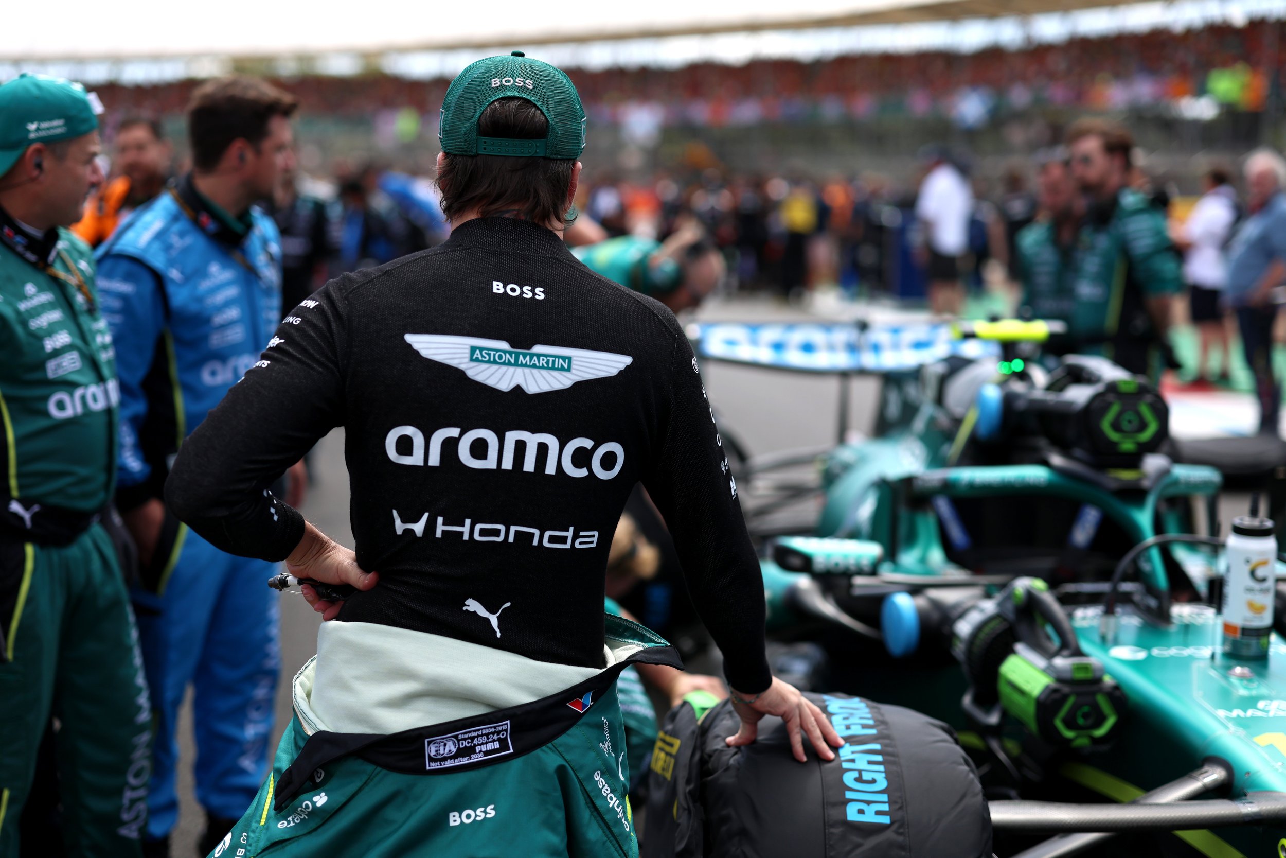 A person in racing gear, with a green cap labeled 'BOSS', stands in the pit area of a racing event, surrounded by other team members and racing equipment, with a blurred crowd in the background.