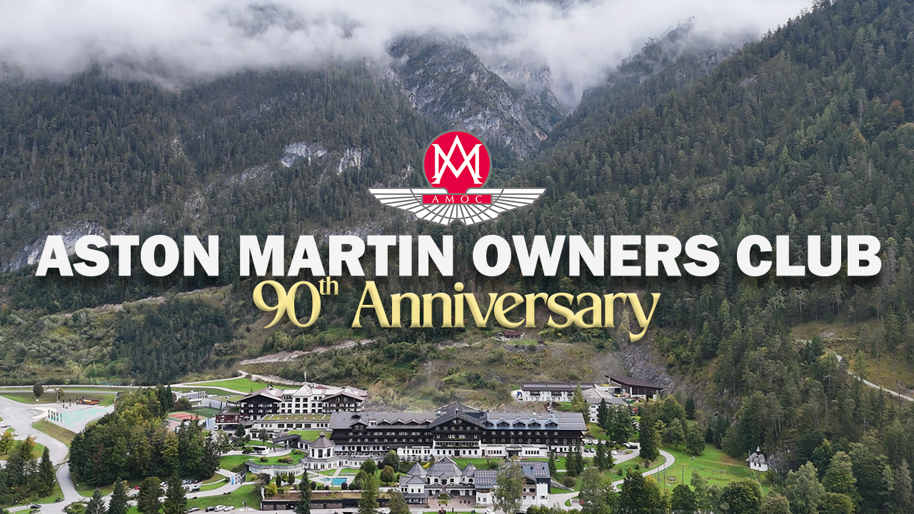 Aerial view of a large mountain resort with lush green trees and buildings, with mountains and cloudy sky in the background. Overlaid text says: 'Aston Martin Owners Club 90th Anniversary.'