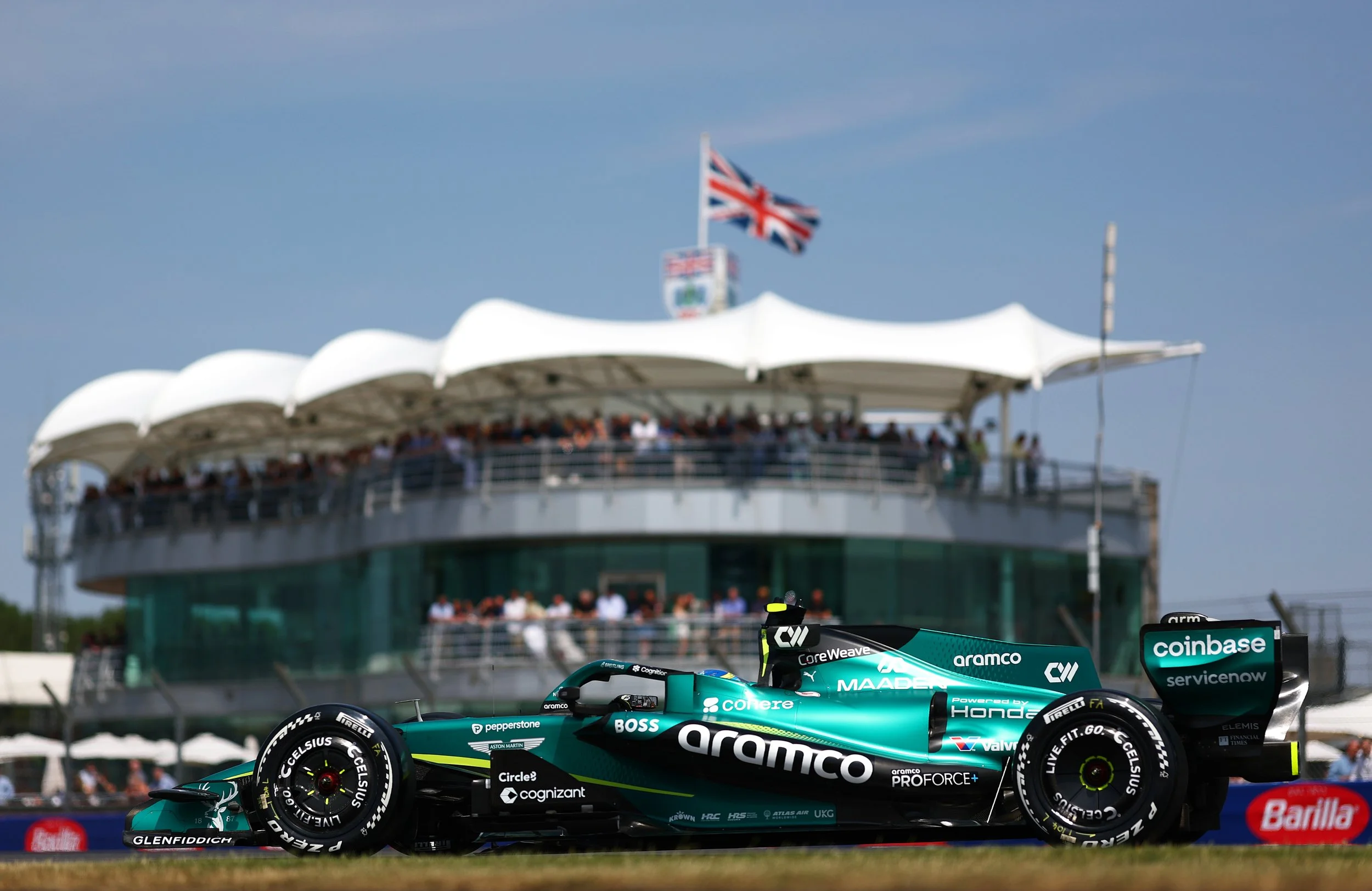 A Formula 1 race car on the track at a racing event, with a grandstand and a British flag in the background.