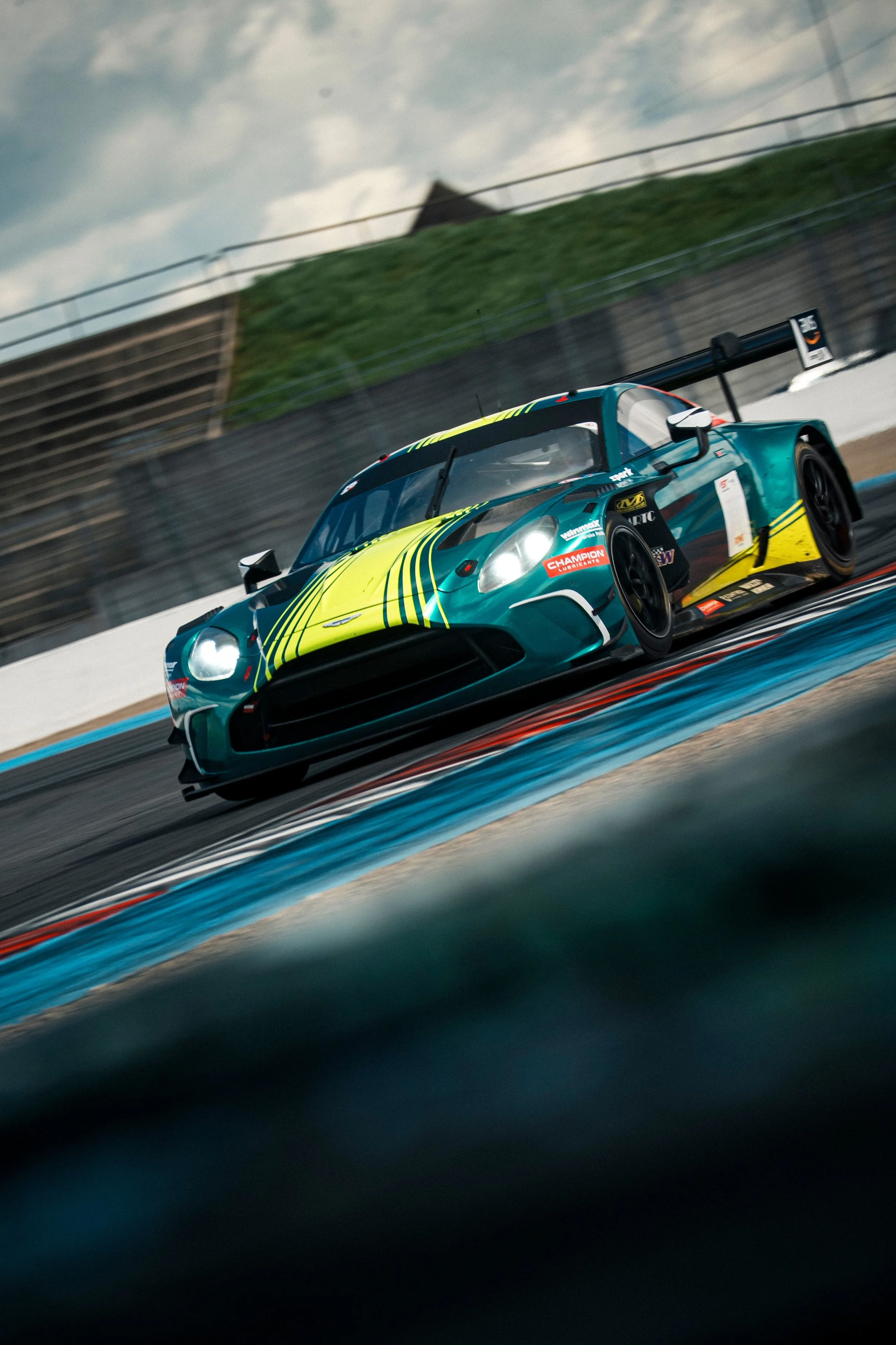 A race car with a teal and yellow livery on a racetrack, blurred to show high speed, with a backdrop of a cloudy sky and green hills.