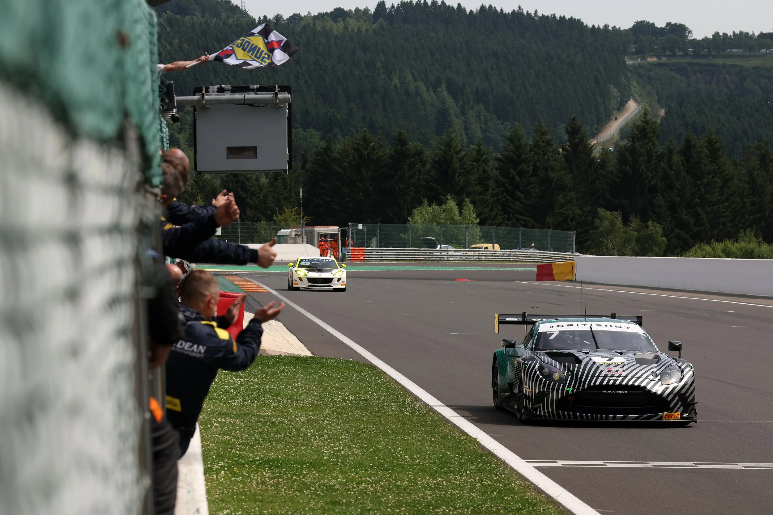 Race cars on a track with spectators watching from the sidelines, surrounded by forested hills and mountains.