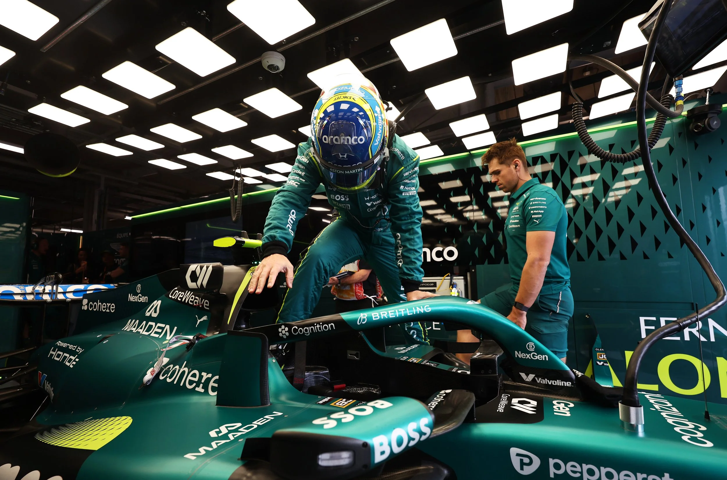 A Formula 1 race car in a garage with two crew members working on it. The driver, wearing a blue helmet, is kneeling in the cockpit. The garage has bright ceiling lights and various sponsorship logos on the car.
