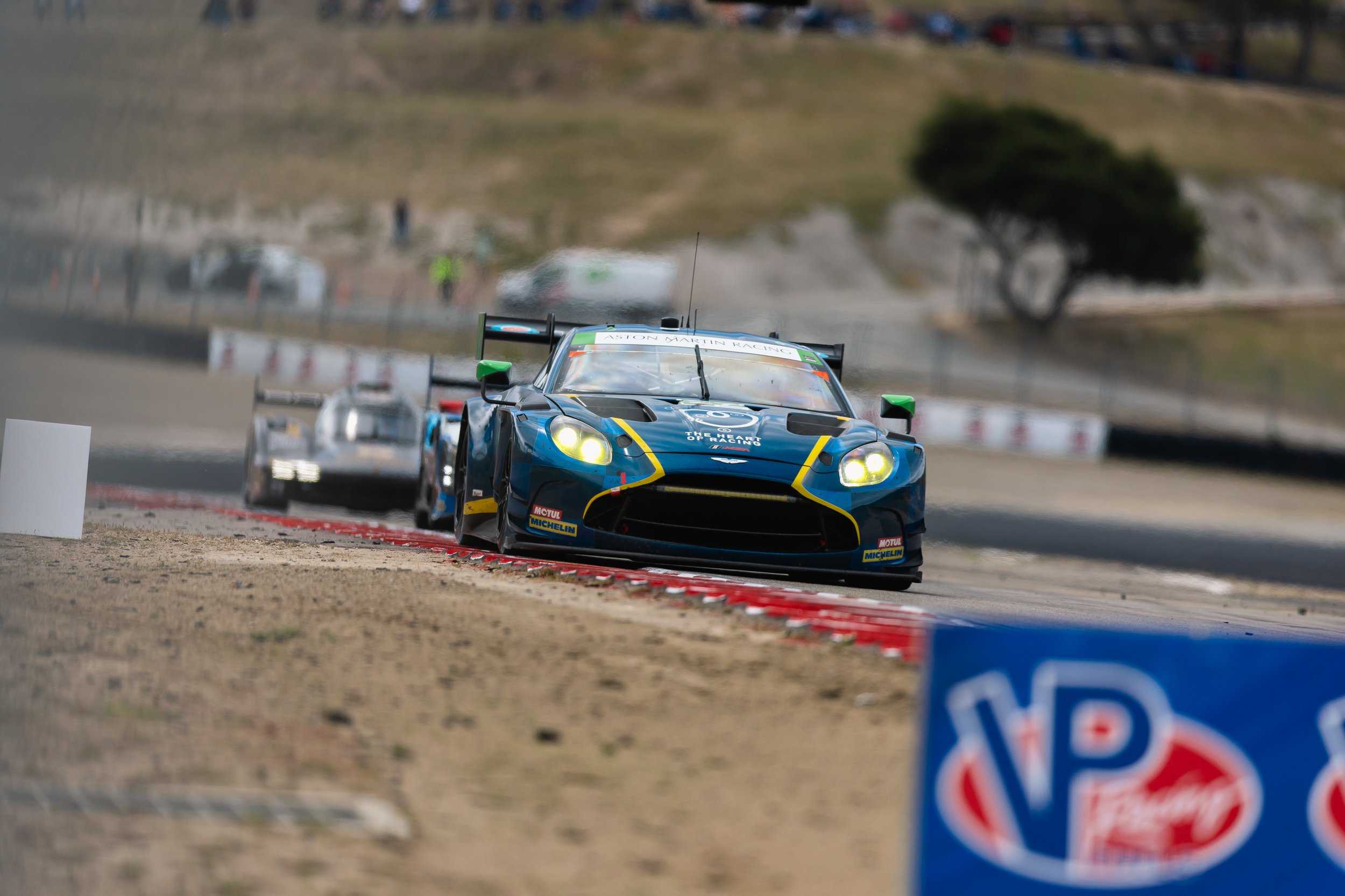 A Heart of Racing Aston Martin Vantage GT3 blue race car with yellow accents on a racetrack during a race, lined with safety barriers and a blurred background of spectators and landscape.