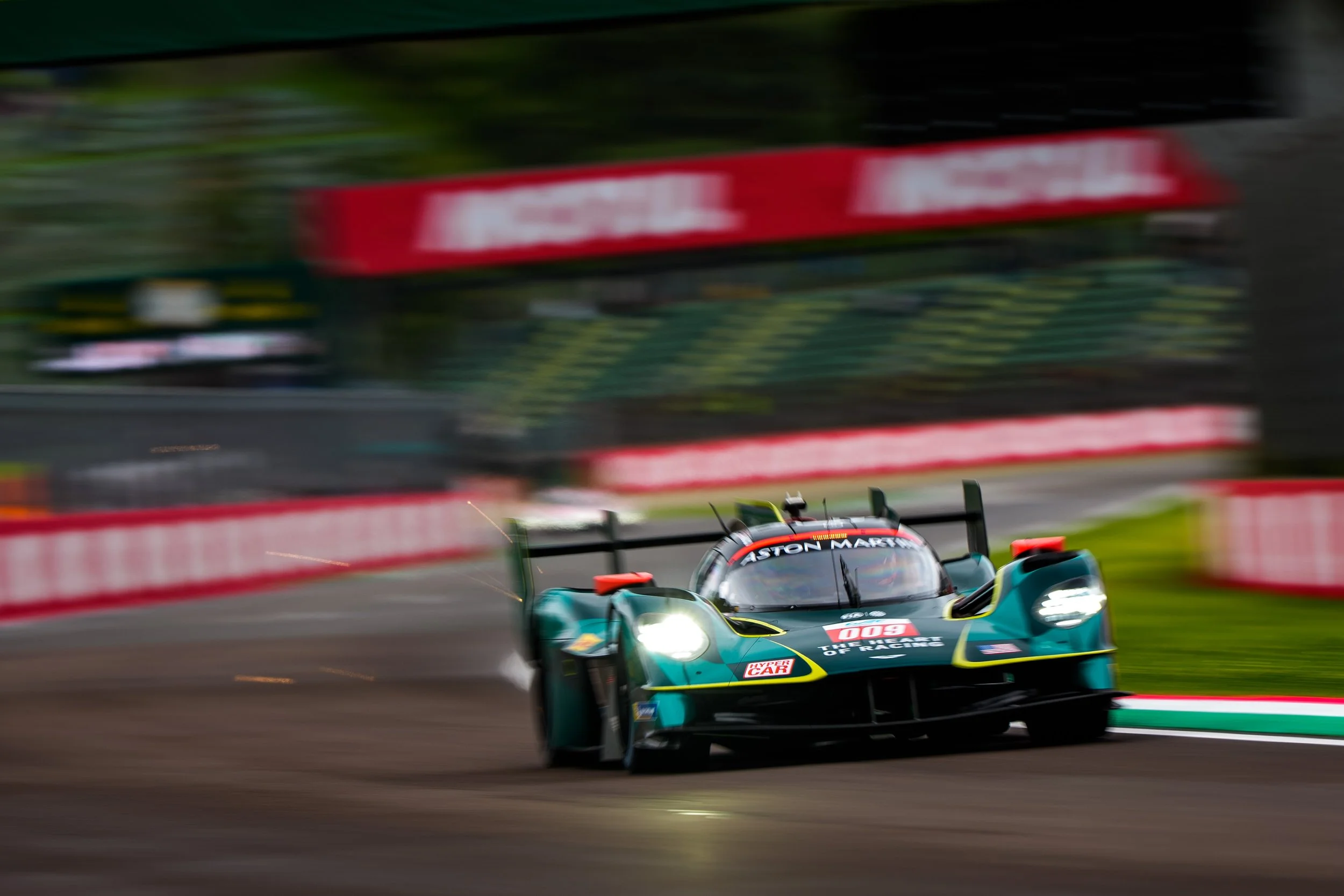 An Aston Martin Valkyrie racing car with the number 009 speeding on a track surrounded by greenery, with motion blur indicating high speed.
