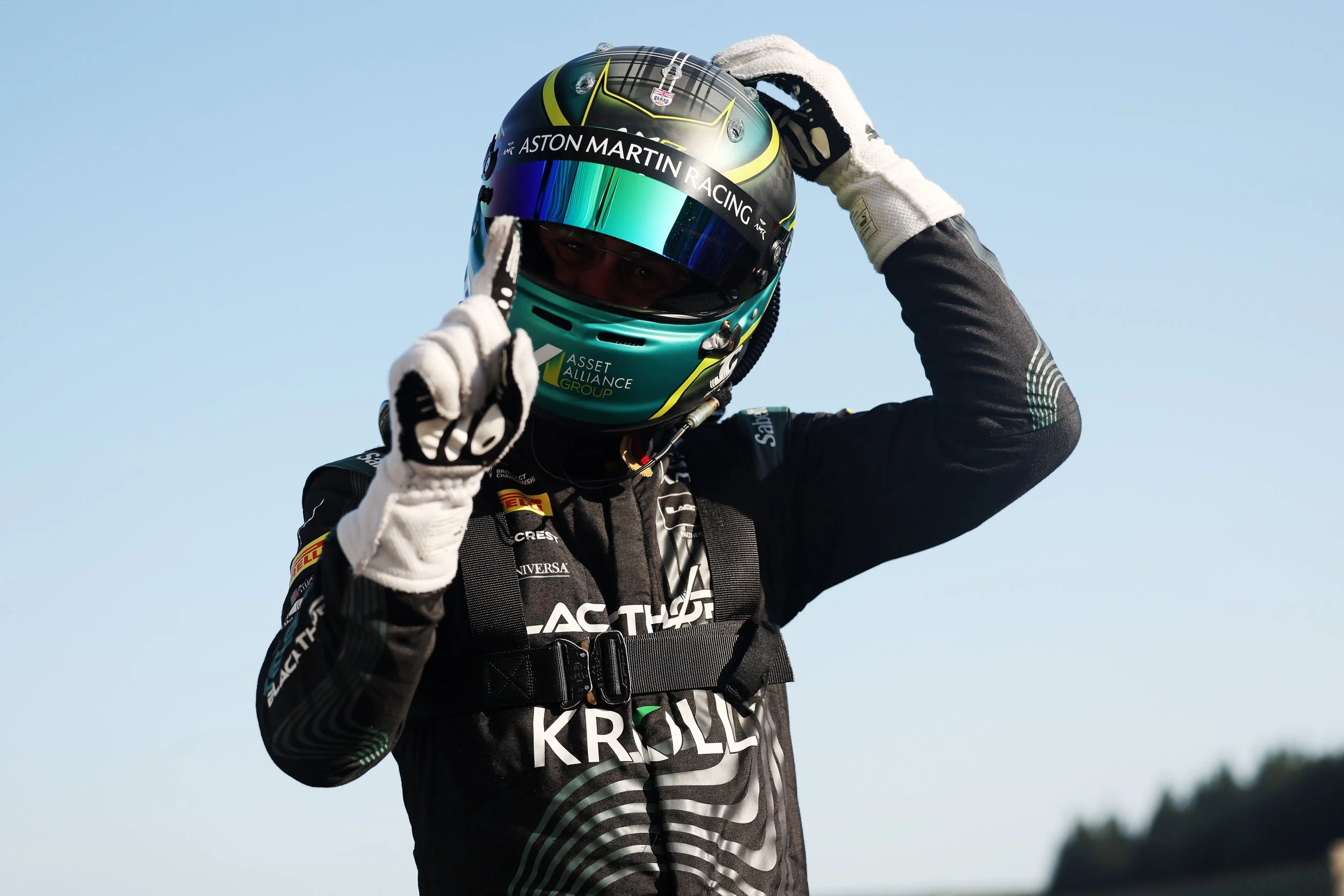 Race car driver wearing a helmet with 'Aston Martin Racing' and 'Asset Alliance Group' logos adjusting the helmet, dressed in black racing suit, standing outdoors against a clear blue sky.