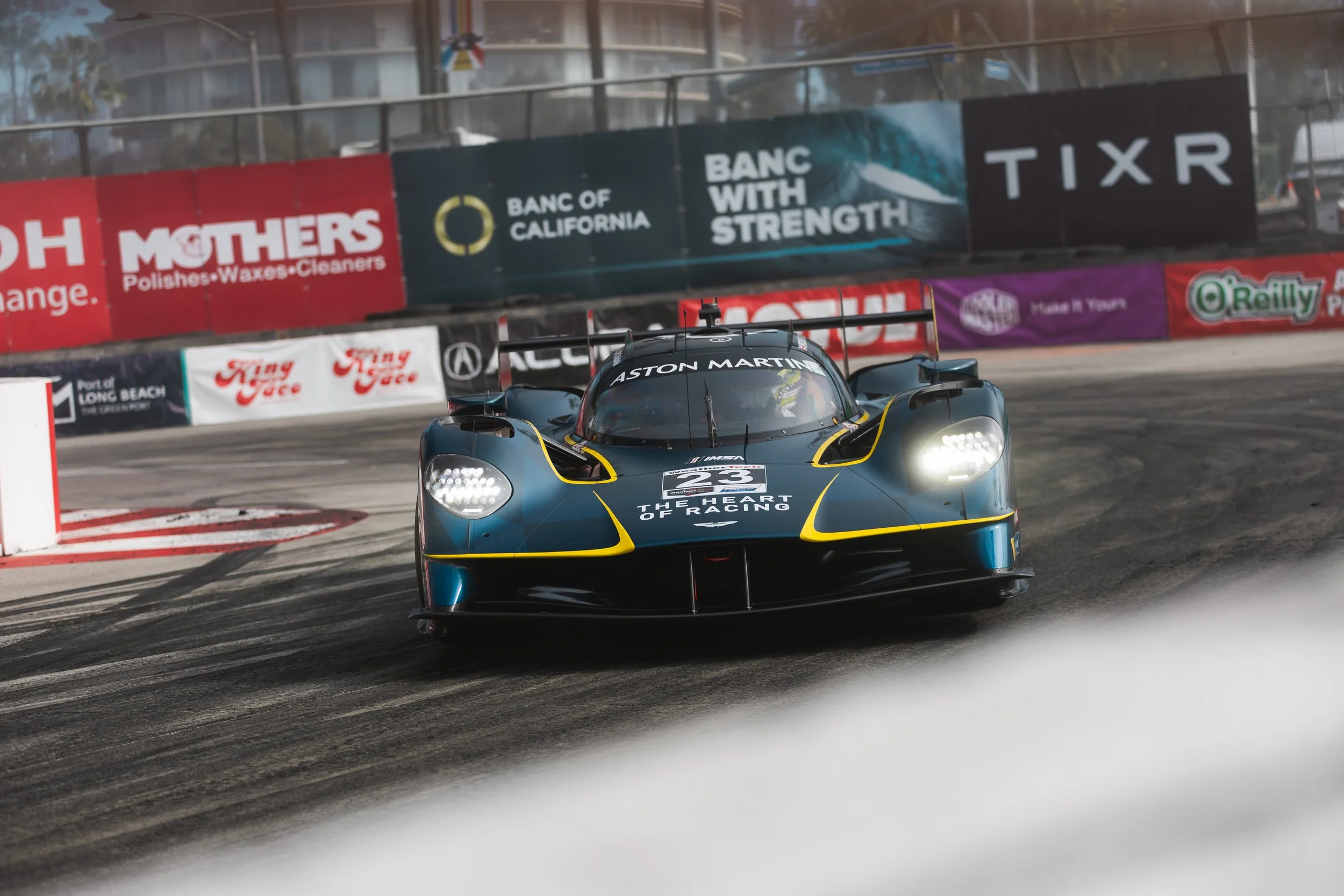A racing car with the number 23 on the front, with Aston Martin branding, speeding around a track with tire marks and barriers adorned with various sponsor banners including Bang & Olufsen, TIXR, and O'Reilly.