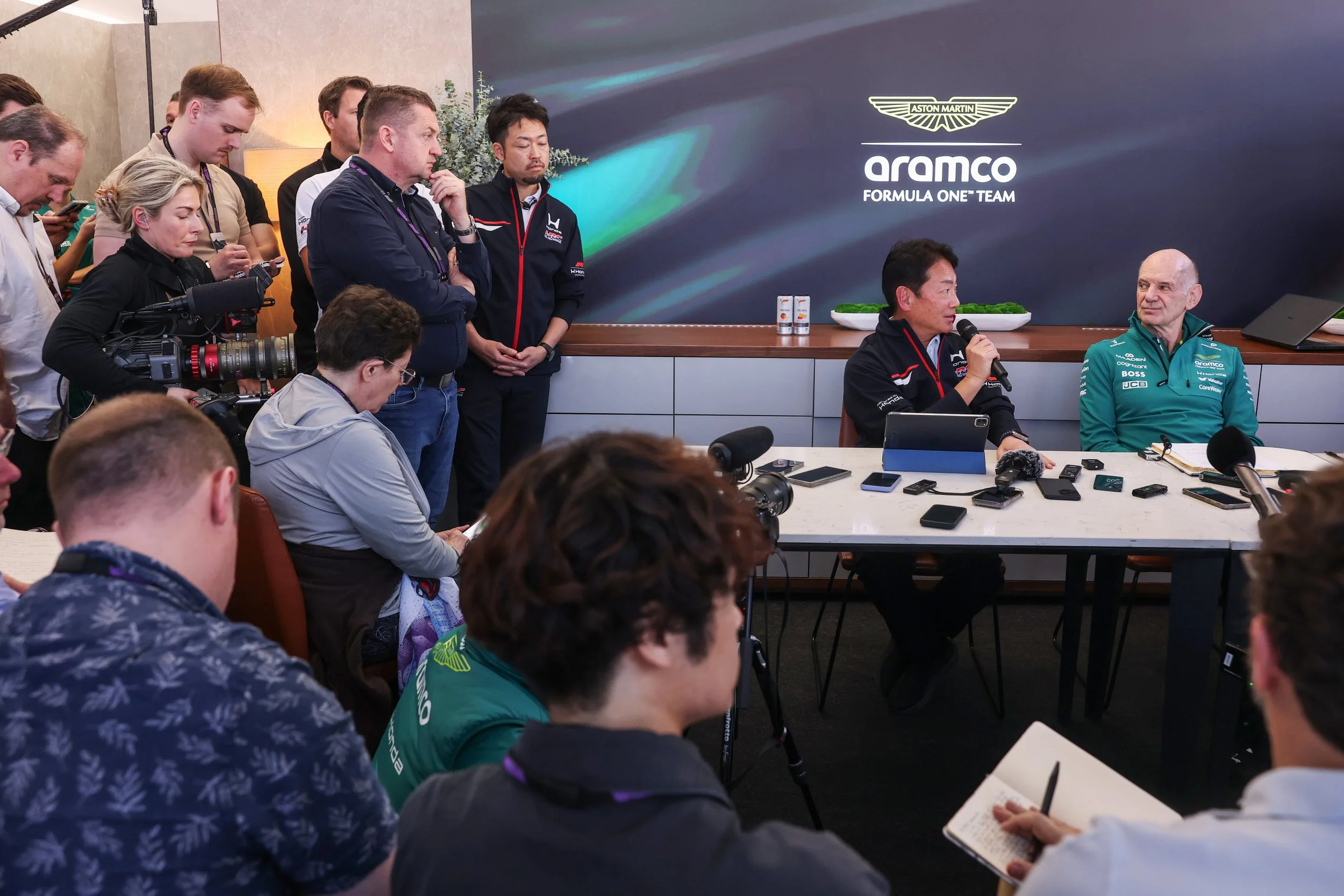 A group of people attending a press conference or meeting, with two men seated at a table, one speaking into a microphone, and a large screen displaying logos for Aston Martin, Aramco, and Formula One Team in the background.