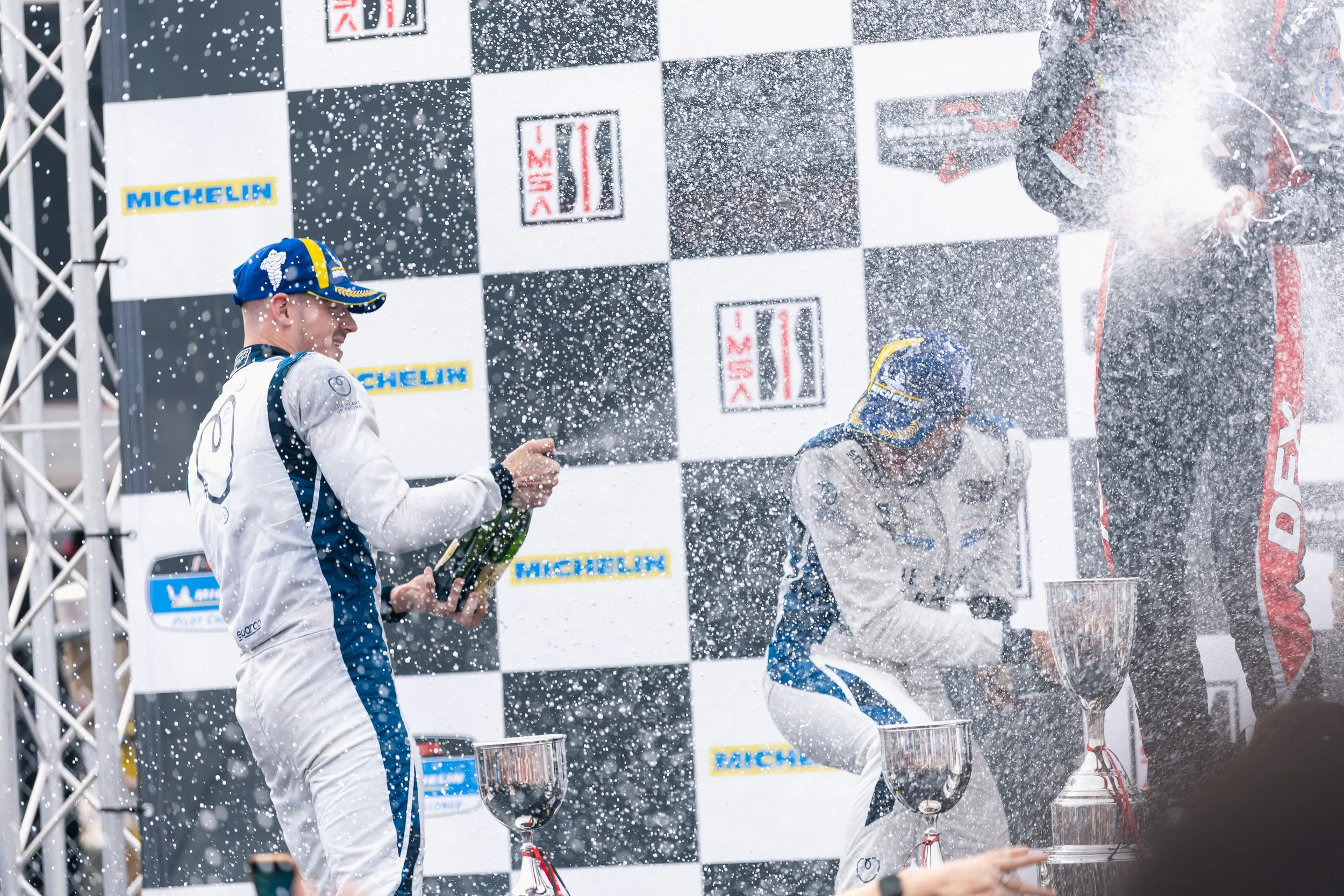 Aston Martin, Heart of Racing Race car drivers celebrating on the podium with champagne, spraying it in the air, after winning a race. One driver is holding a bottle of champagne, and others are standing near trophies on a checkered flag background.