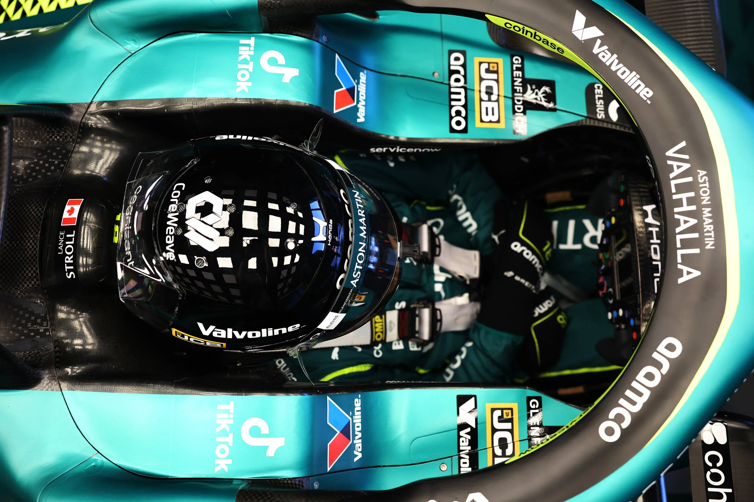 Left side view of a Formula 1 racing car driver in a black helmet with a checkered pattern and a reflective visor, sitting inside a teal and black racing car with various sponsor logos including Valvoline, JCB, and Vail Hallast. The image shows the driver's seat, steering wheel, and part of the car's cockpit.