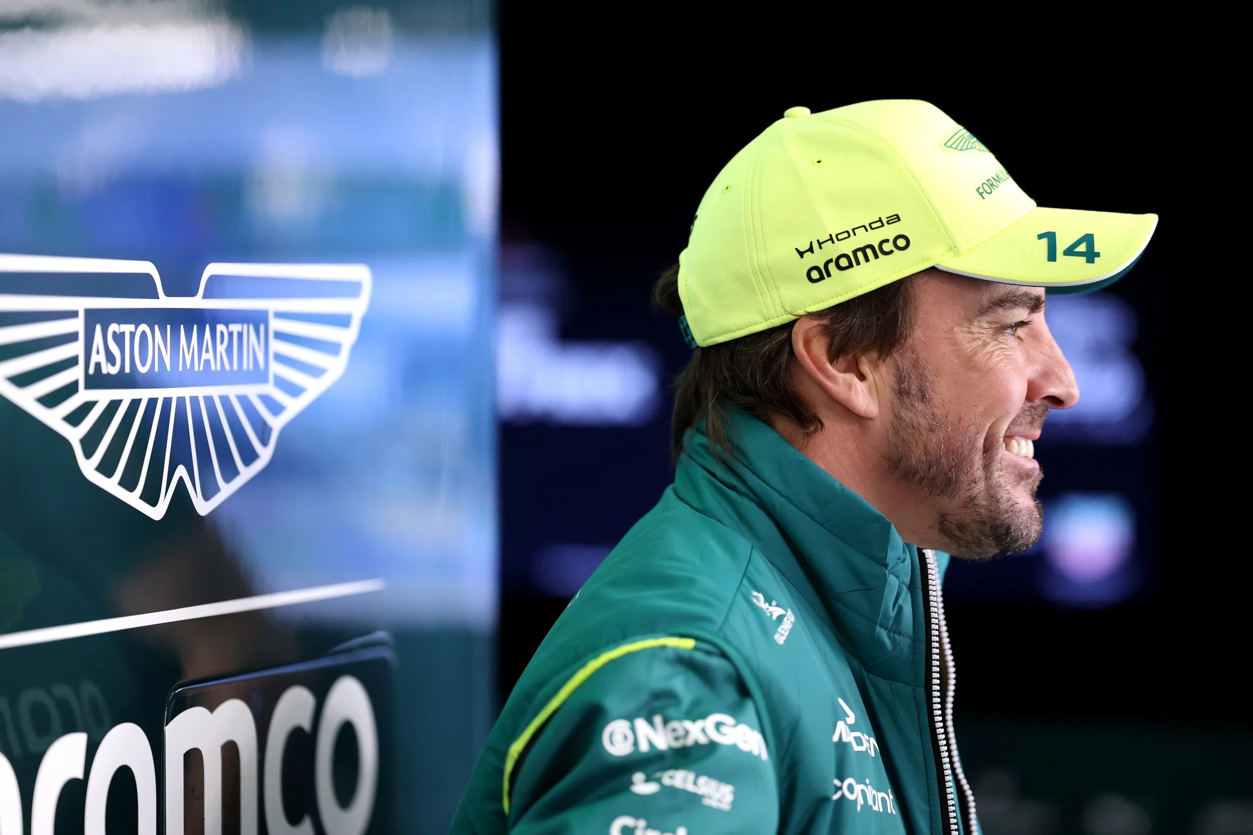 A man wearing a bright yellow cap with the words 'Honda Aramco' and the number 14 on it, smiling, in a racing suit with various sponsor logos, standing near a blue sign with the Aston Martin logo.