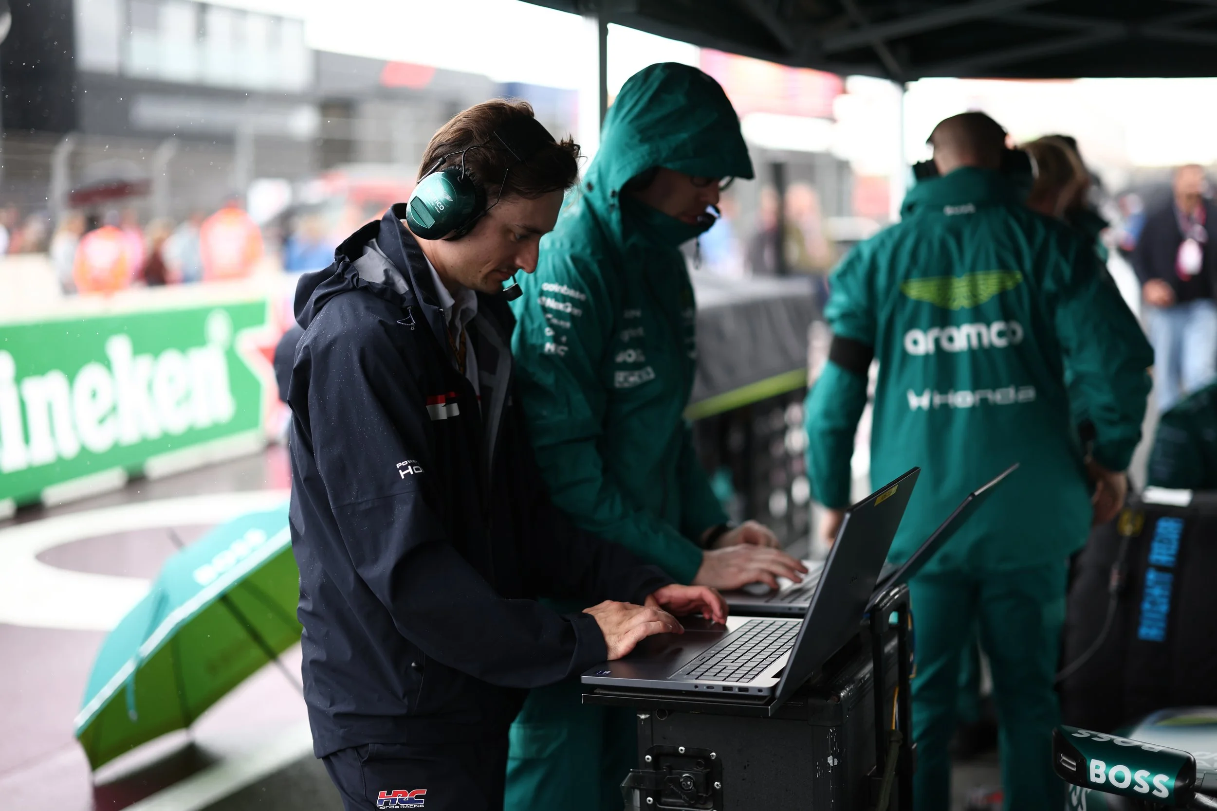 Two men and a woman working on laptops under a tent at a racing event. The man on the left is wearing a black jacket with a headset. The woman and man on the right are wearing green racing jackets with logos. The background shows blurred spectators and a green Heineken banner.