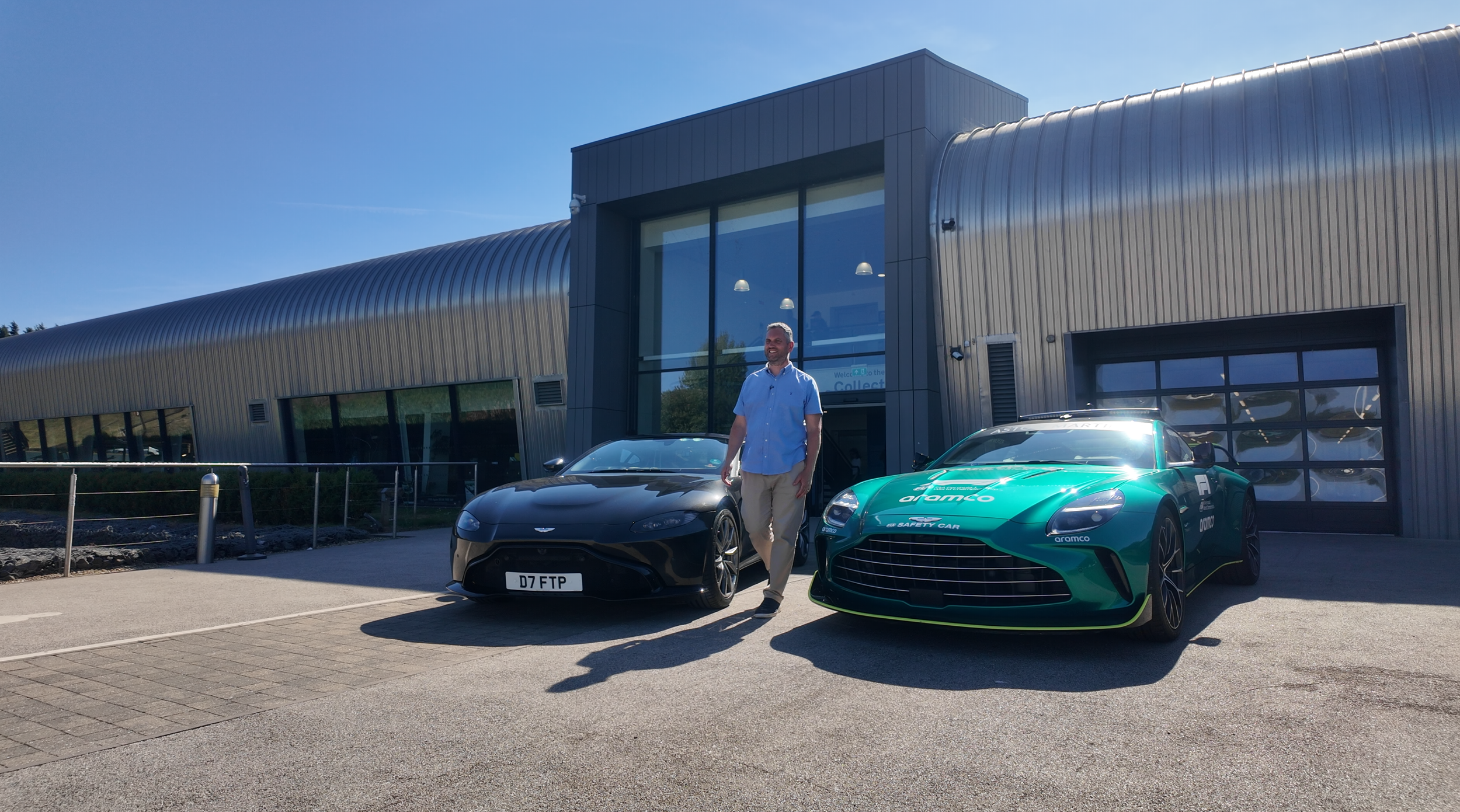 Dan from Fuel the Passion, walking between two sports cars, a black Aston Martin and a green racing car, in front of a modern industrial building on a sunny day.