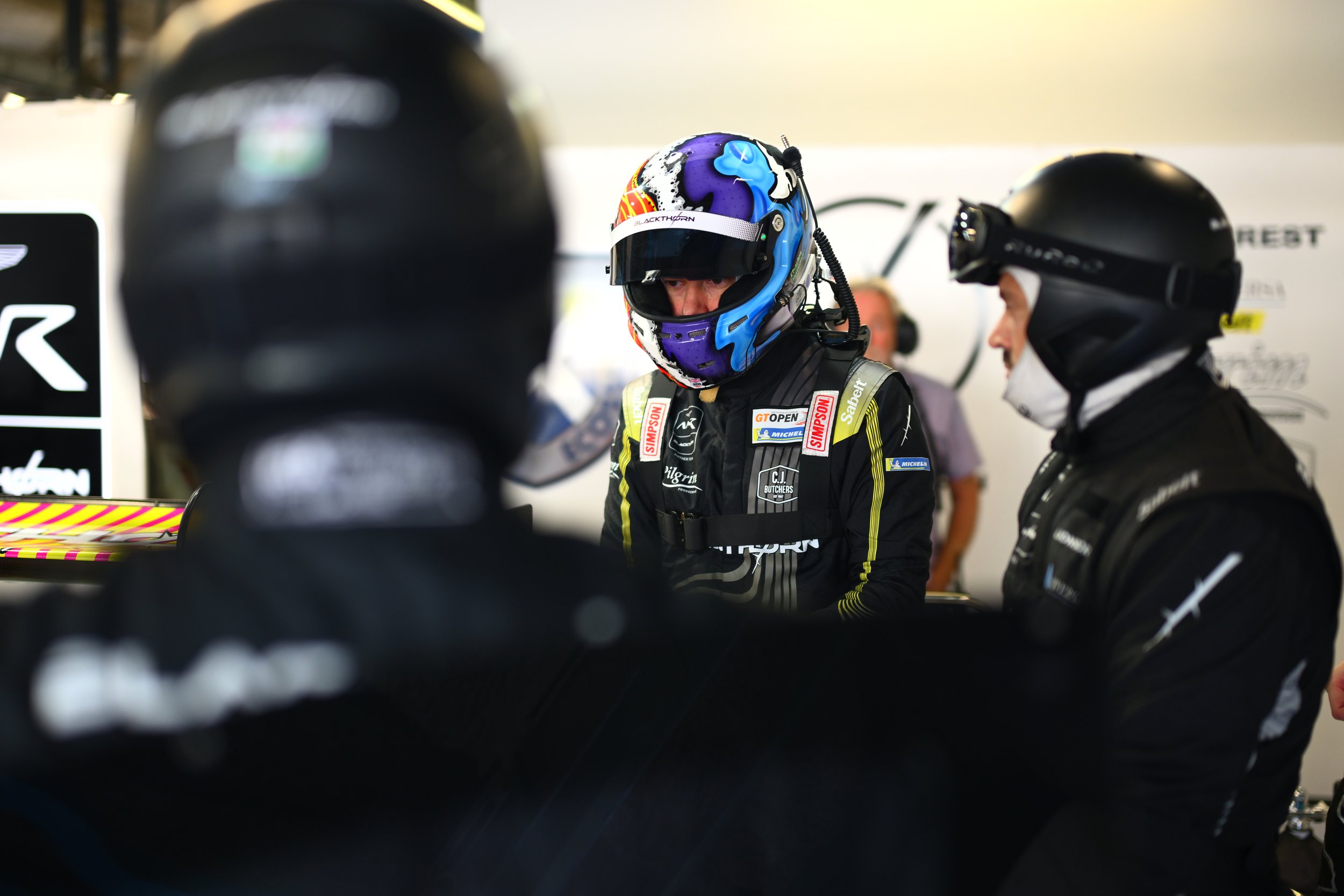 Race car driver in a racing suit and helmet in the garage, surrounded by team members.