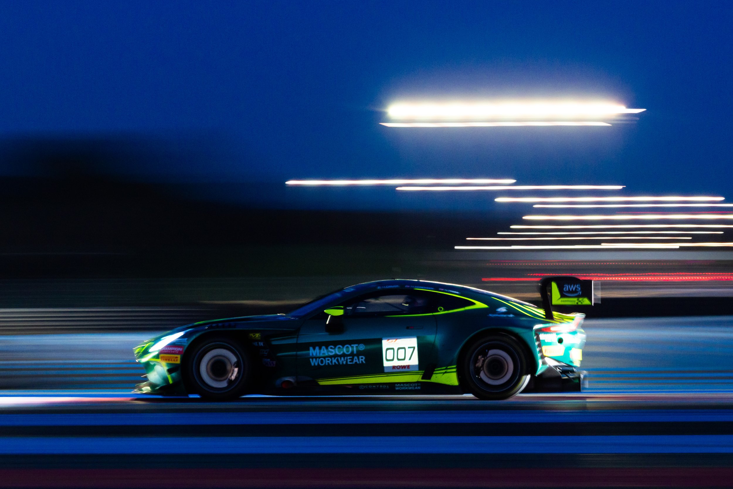 A racing car with sponsorship decals moving at high speed, blurred lights in the background indicating motion, night time setting.
